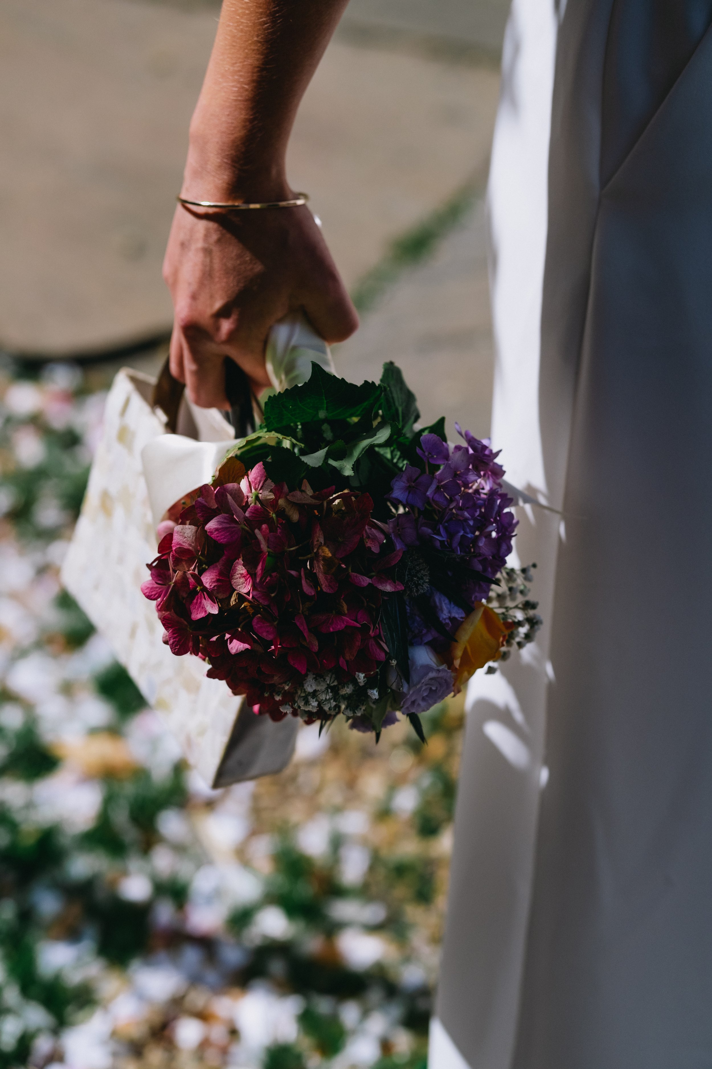 A person dressed in a white suit with a gold bracelet on their wrist is holding a bouquet of purple and pink flowers. The background appears to be an outdoor setting with blurred greenery.