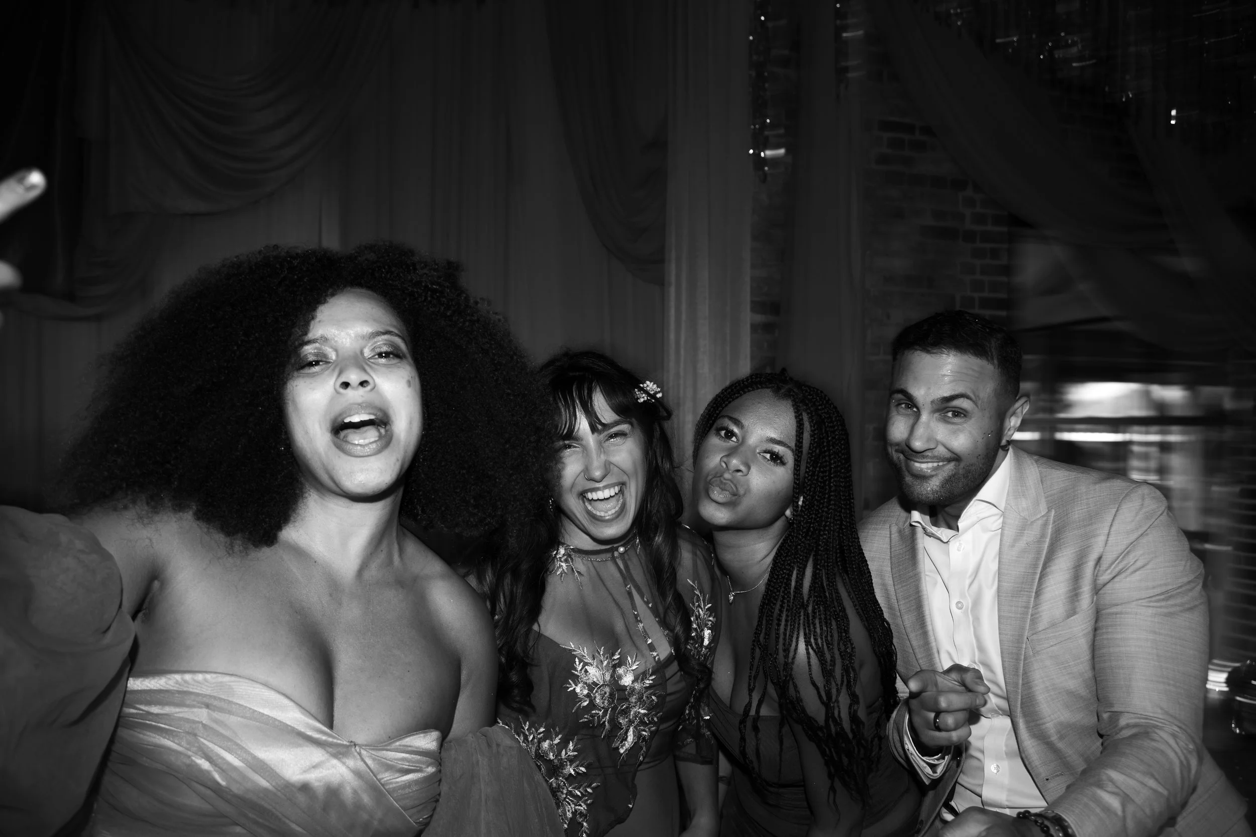 A group of five diverse people posing for a photo at a social event, with a woman in the foreground taking a selfie. They are smiling and dressed in semi-formal attire, with curtains and a brick wall in the background.
