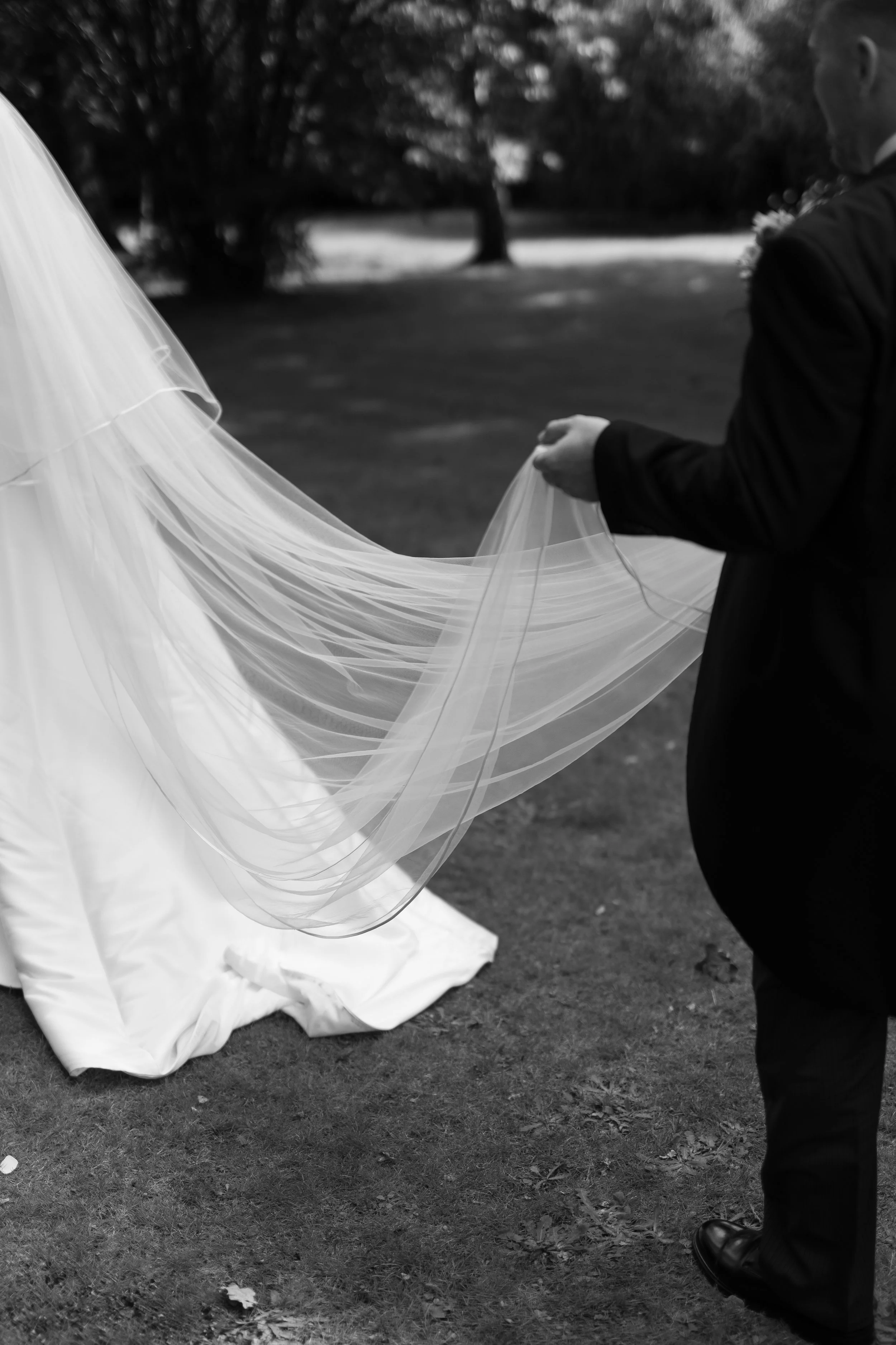 A person dressed in a dark suit, possibly at a wedding, holding a long, flowing piece of fabric or veil outdoors on a grassy area. The person's face is turned away and the background features trees and a pathway.