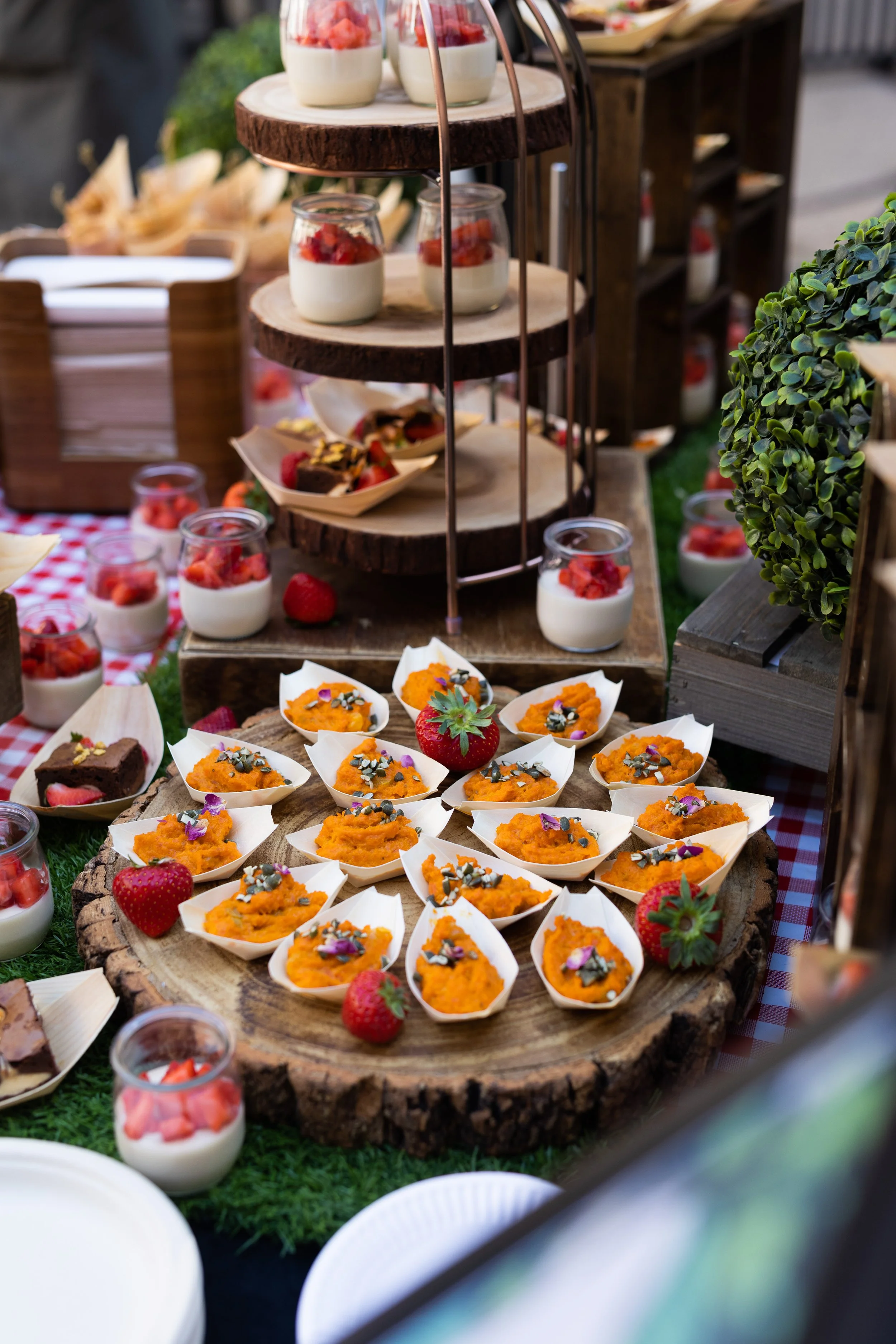 Dessert display featuring small pumpkin-shaped desserts topped with flower petals, surrounded by strawberries and panna cotta cups with strawberries on top, arranged on a wooden slice serving platter at an outdoor event.