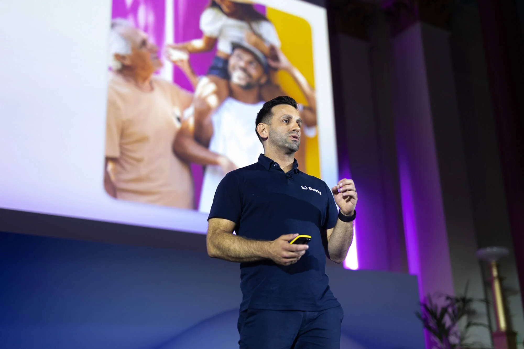 A man is giving a presentation on stage, holding a remote in his right hand. Behind him is a large screen displaying an out-of-focus image of people, including a man and a woman, engaging in an activity with bright purple and yellow background colors.