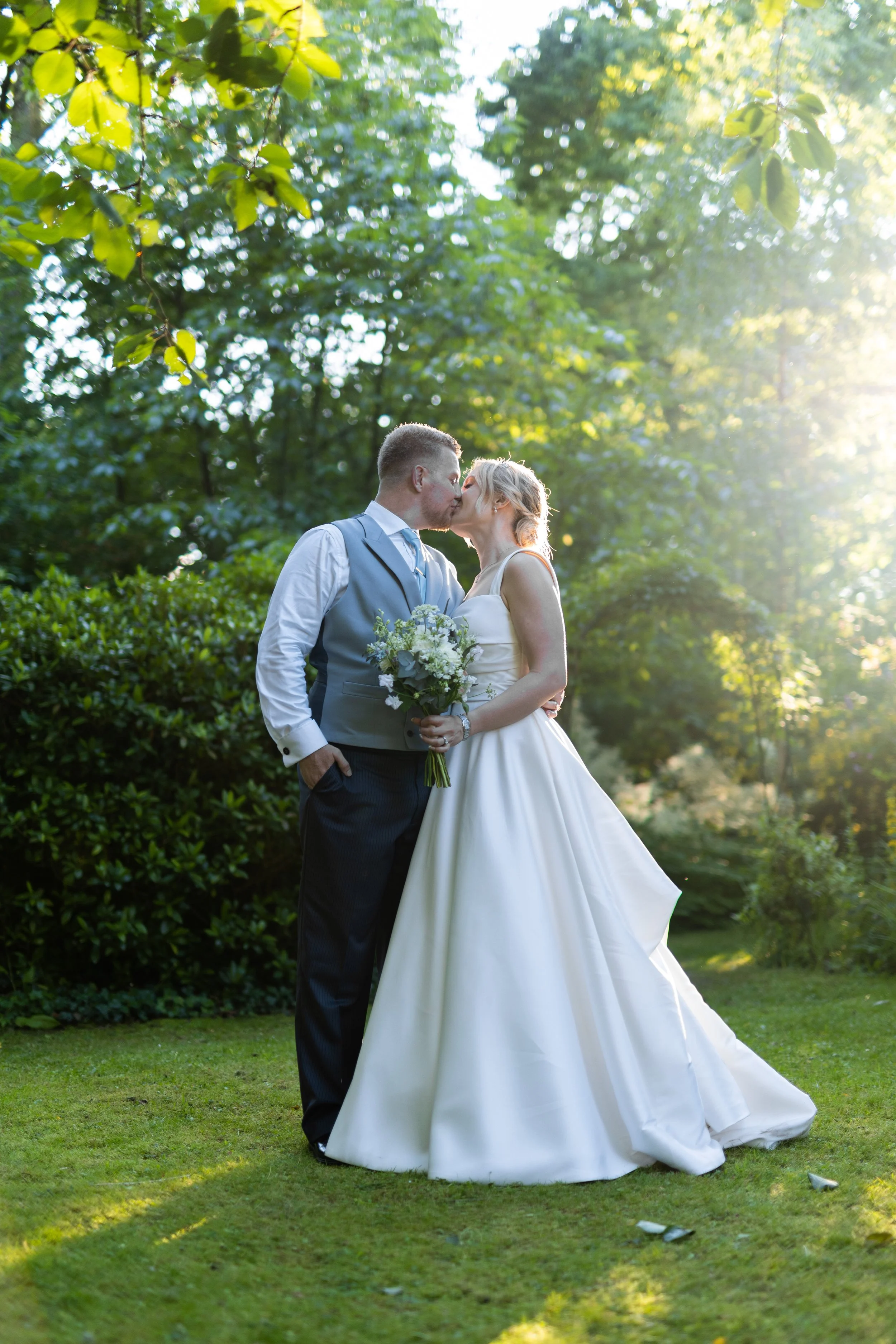 A bride and groom share a kiss outdoors in a lush garden during daytime, with sunlight filtering through trees behind them.