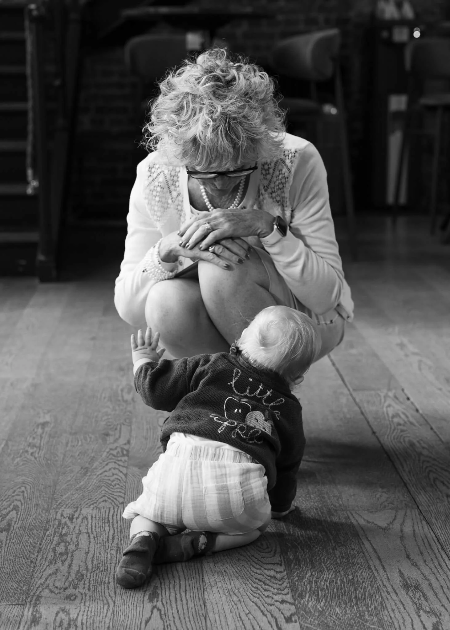 An elderly woman with curly hair, glasses, and wearing a pearl necklace, sitting on the floor. She has her hands clasped and is looking down. A young child is kneeling in front of her, reaching towards her. The setting appears to be indoors with chai