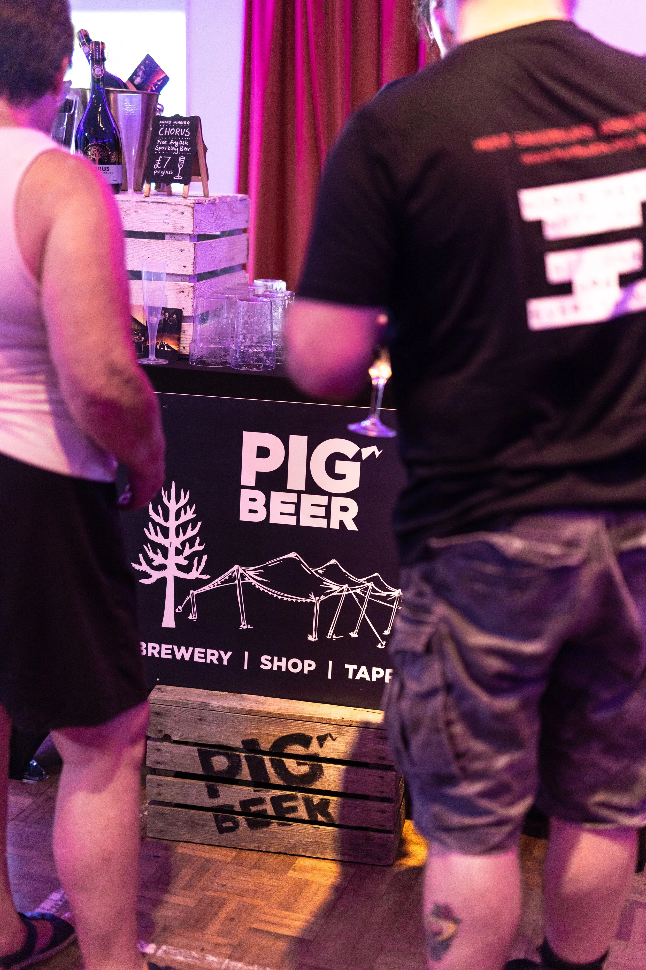 People standing around a bar area at a brewery or taproom, with a wooden crate bar displaying drinks and a sign advertising a sparkling beer for £7 per glass. The background has purple lighting and red curtains.