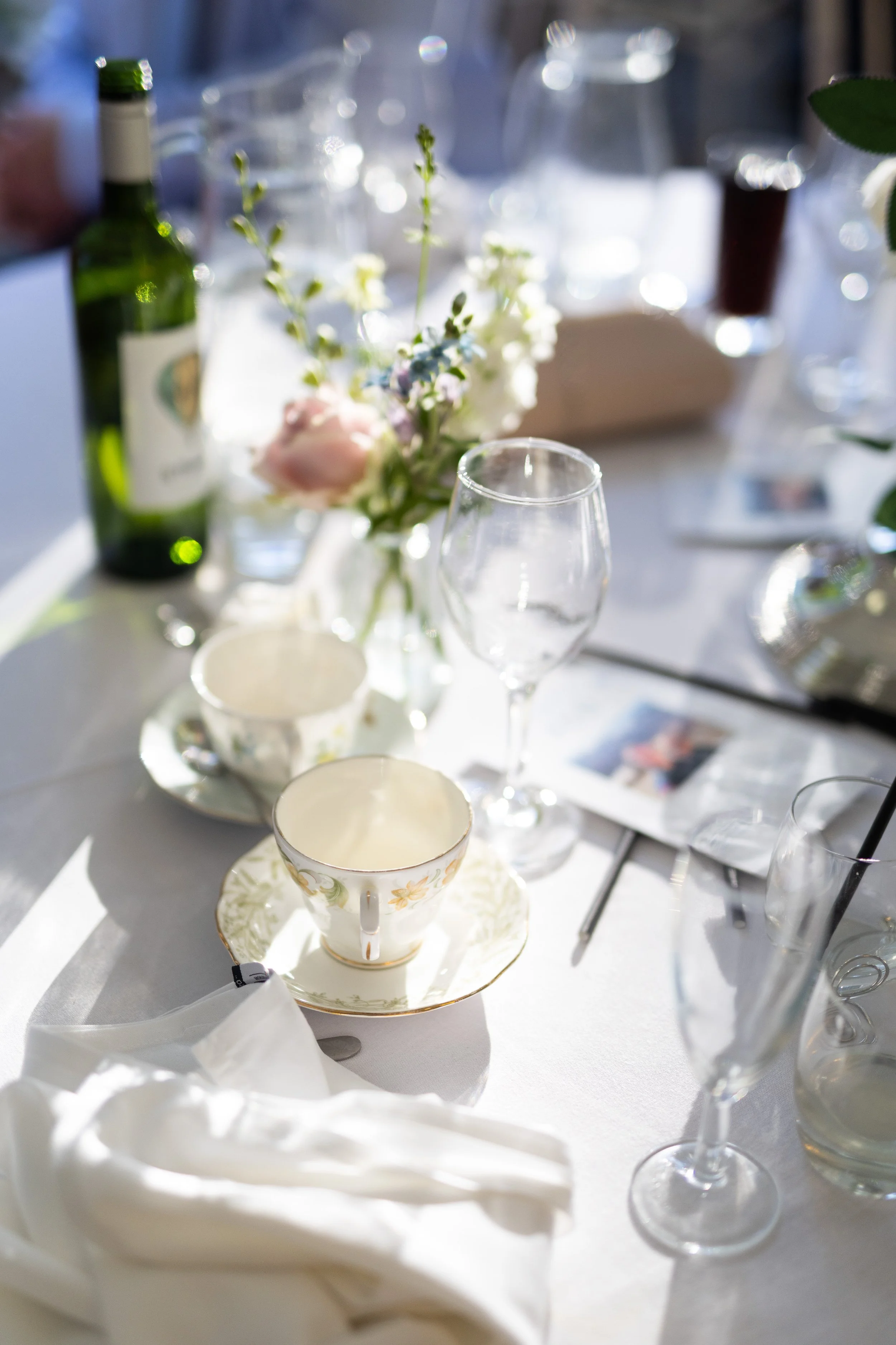 A table set for a meal with a flower arrangement, wine bottles, wine glasses, and tea cups with saucers, all on a white tablecloth.