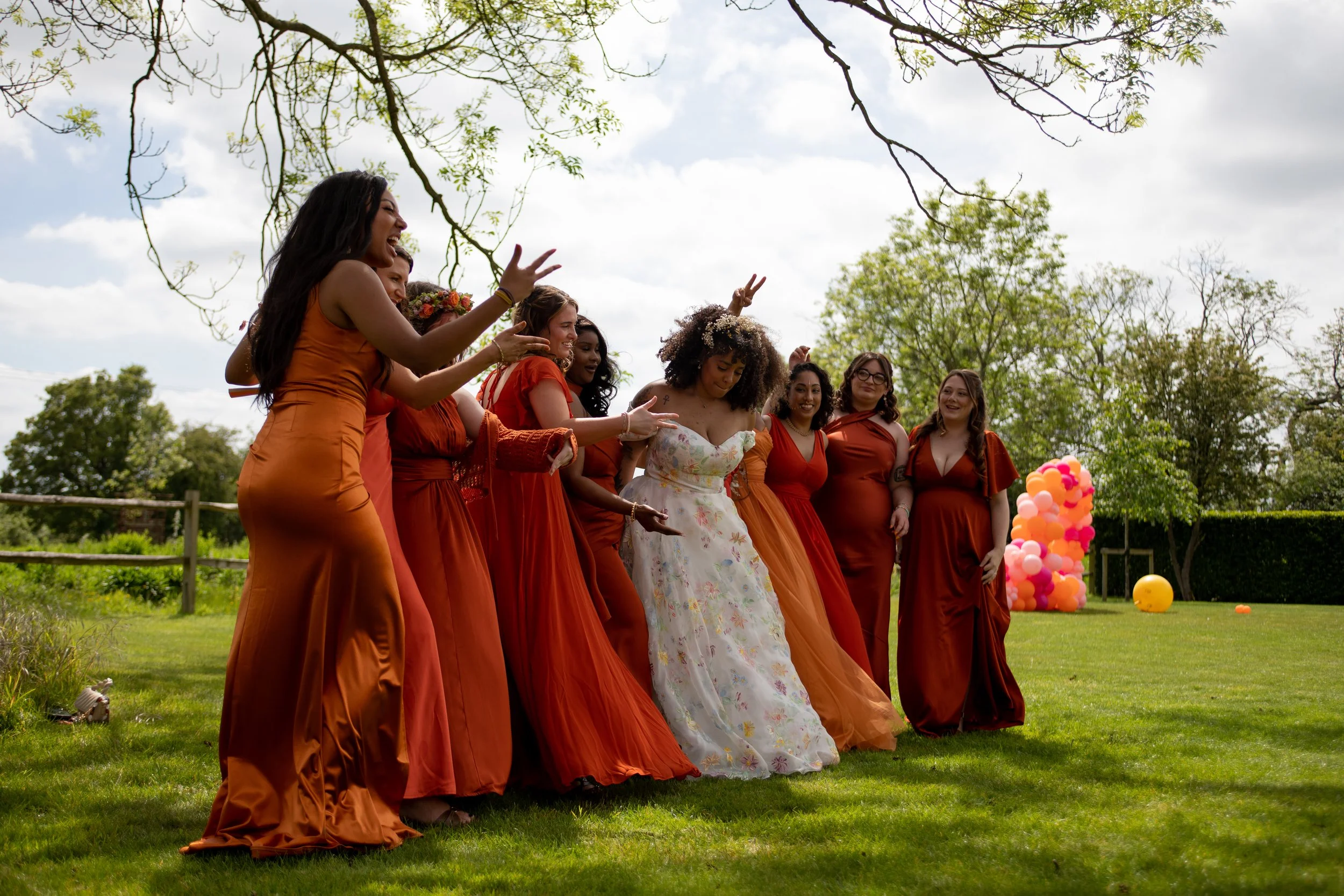 A group of women in orange and floral dresses standing outdoors on grass, celebrating with balloons and smiling under partly cloudy skies and green trees.