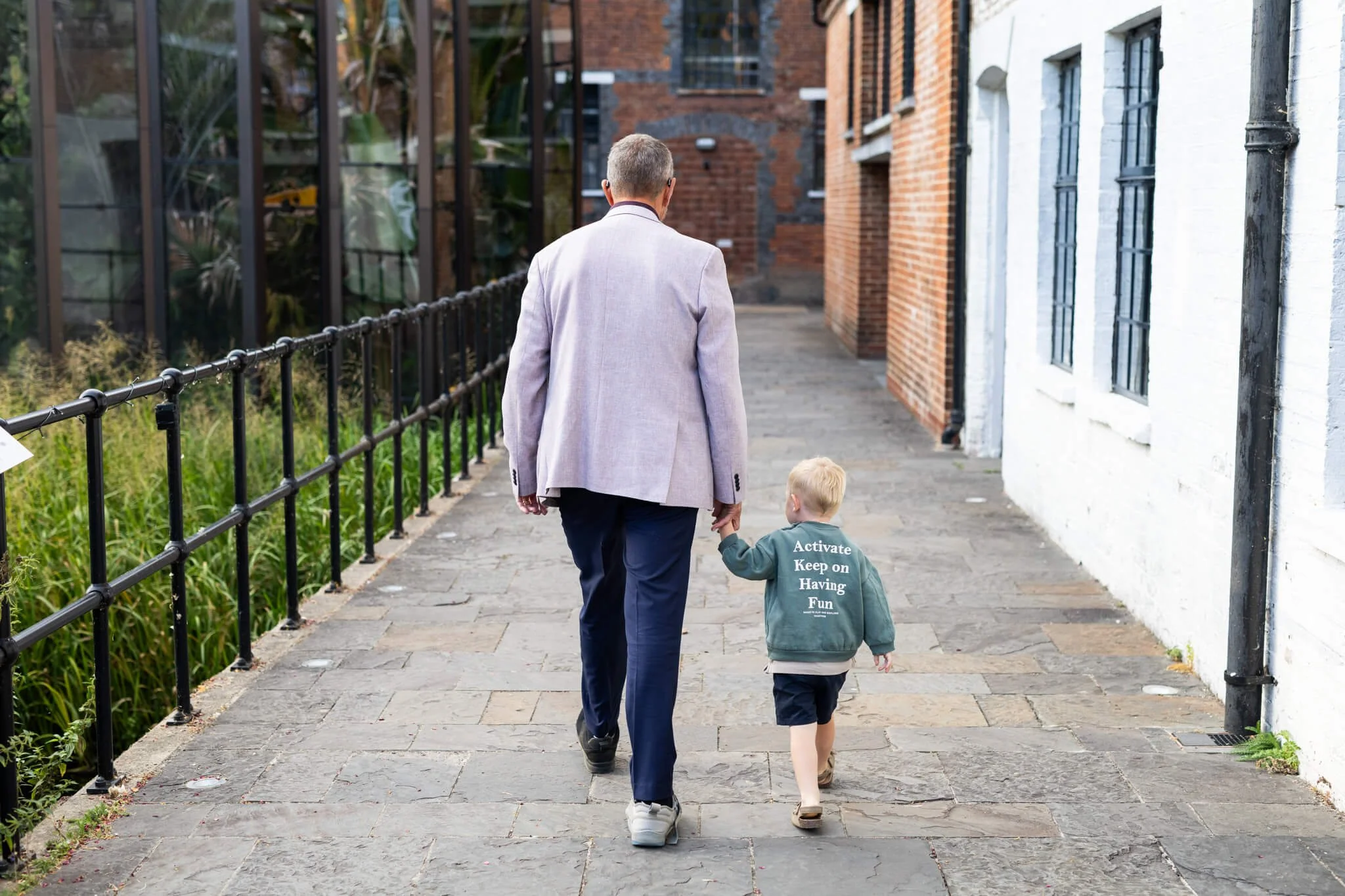 A man in a light gray blazer holding hands with a young boy walking along a city sidewalk with brick and white buildings.