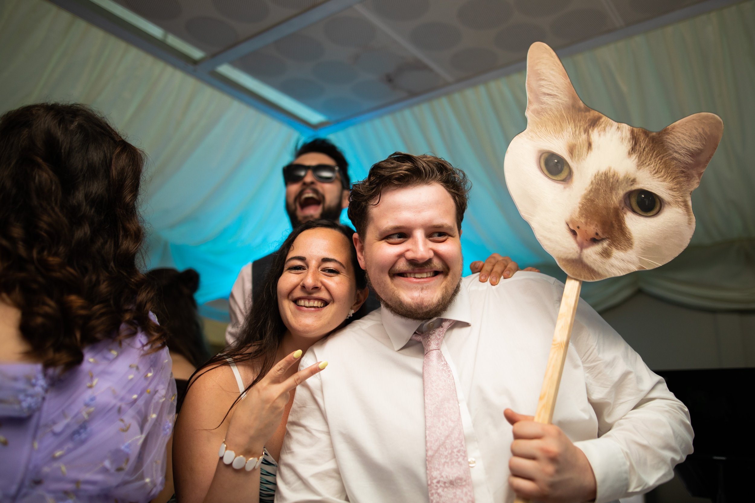 Group of people smiling and taking a selfie, one person holding a cat-shaped mask on a stick.