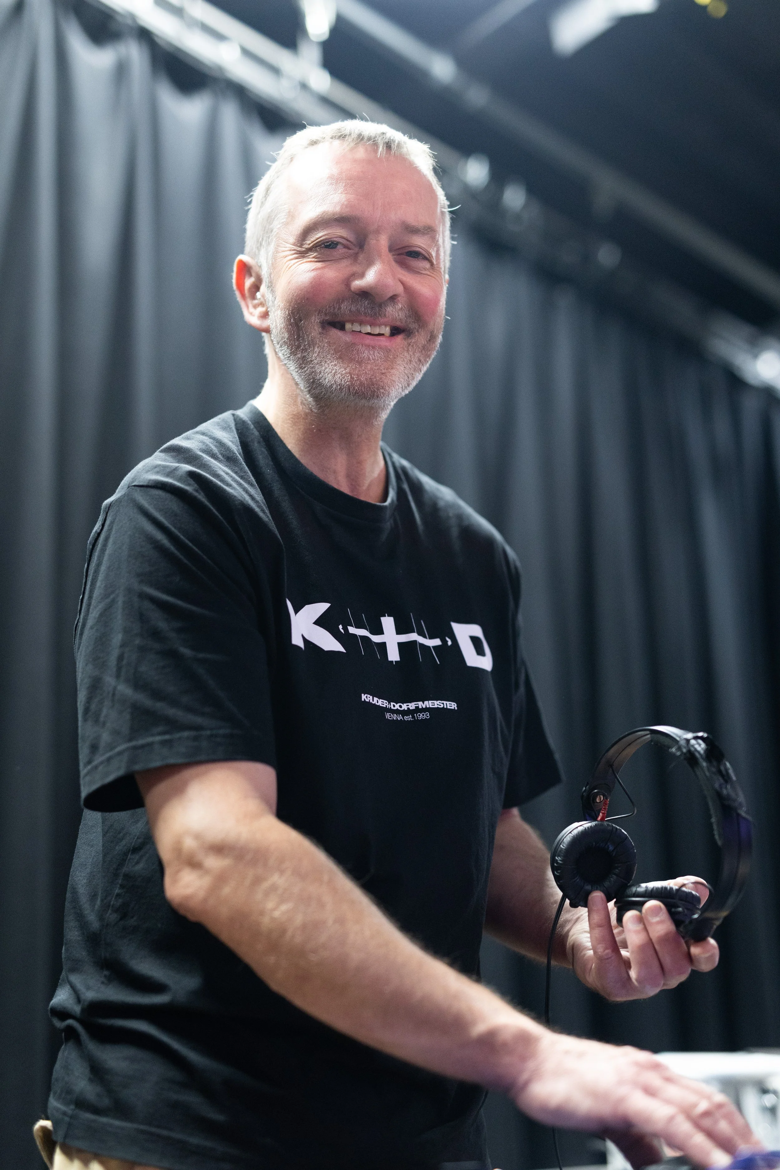 A smiling middle-aged man with gray hair and beard holding headphones in one hand and playing a keyboard or piano with the other, standing in front of black curtains, wearing a black T-shirt.