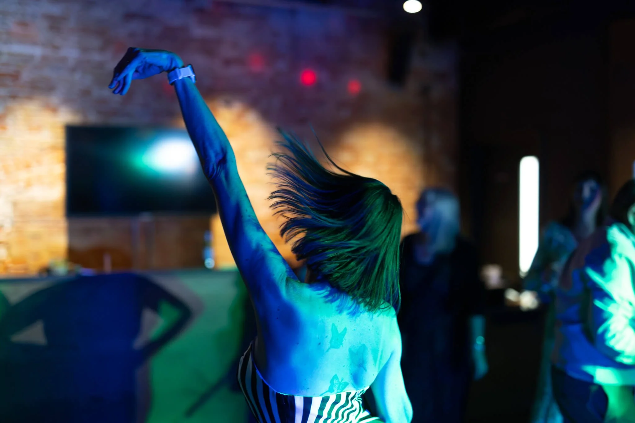 A person with long hair painted in neon colors, dancing in a dark room with vibrant lighting and an exposed brick wall in the background.