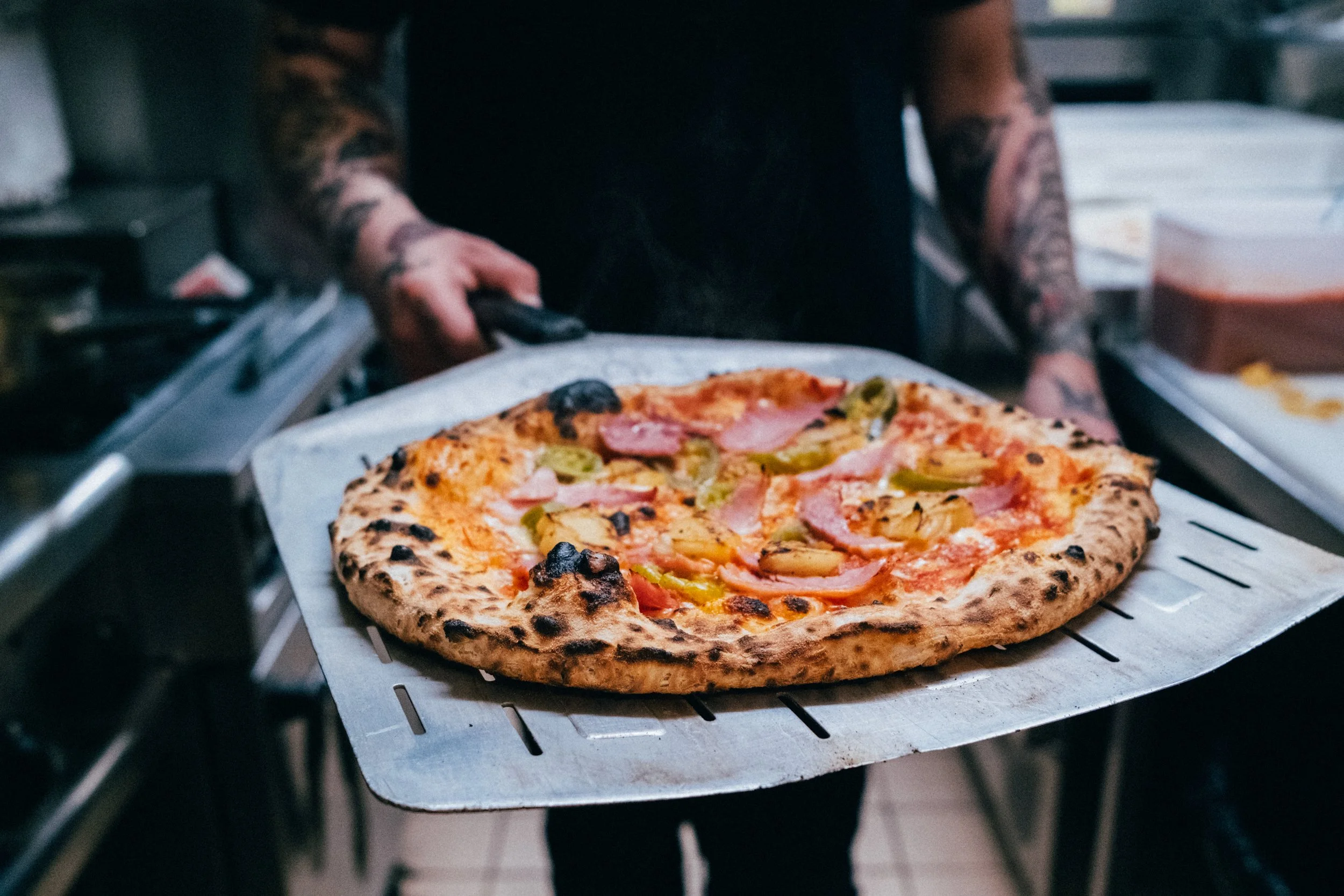 A pizza with toppings including ham, pineapple, green peppers, and black olives being removed from an oven by a person with tattoos.