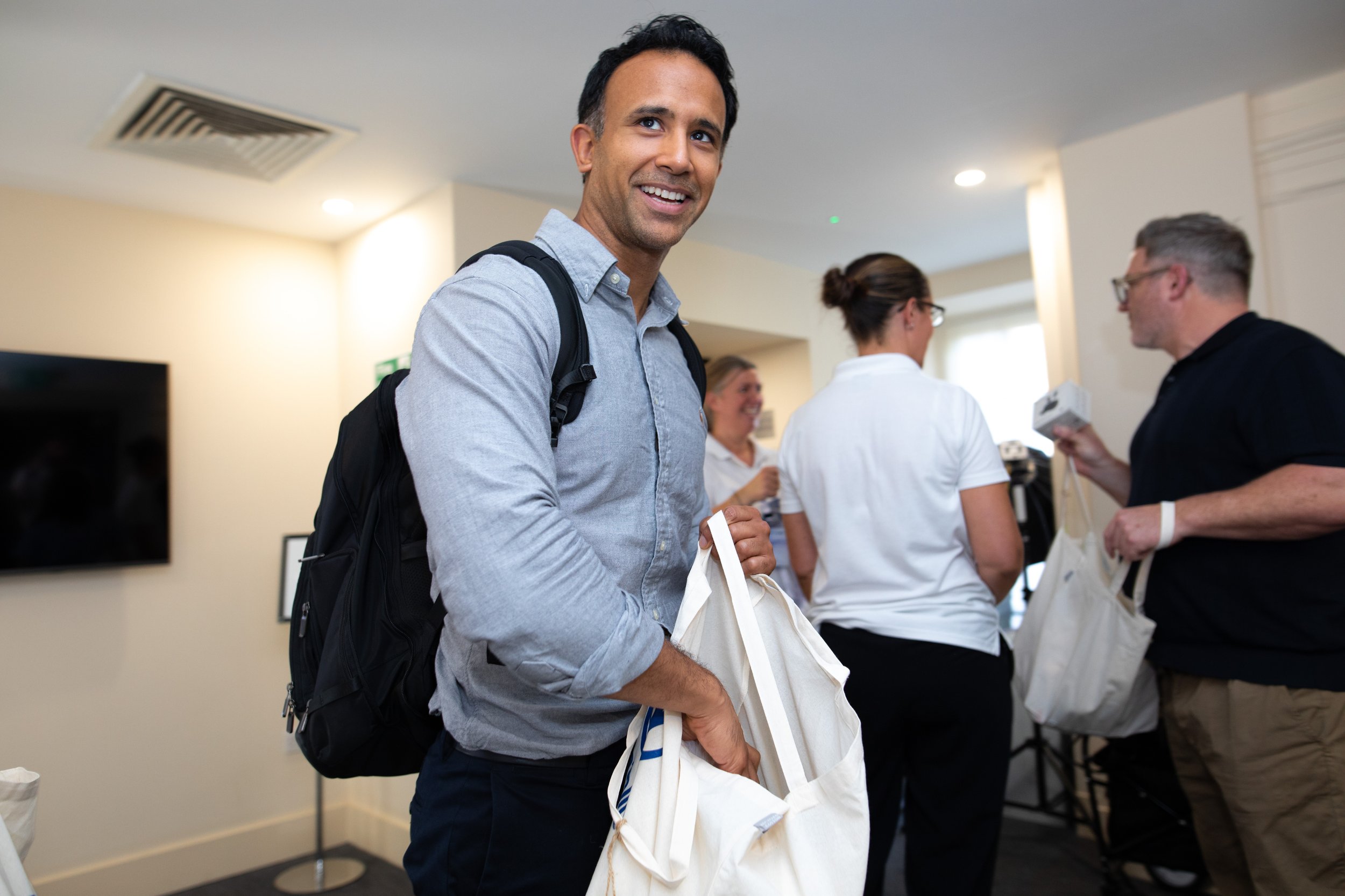 A man with a backpack smiling at an indoor event, holding a tote bag. Several people in the background are engaged in conversations.