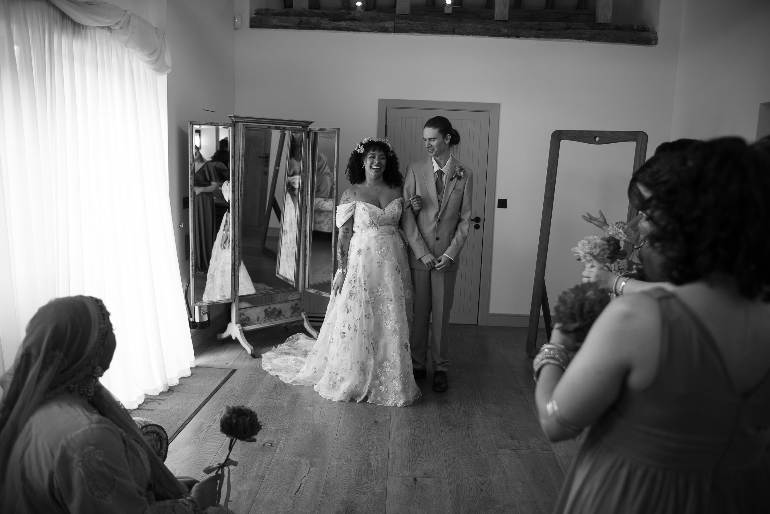 A bride and groom standing together, smiling happily, at a wedding ceremony in a room with wooden accents and mirrors, with seated guests observing.