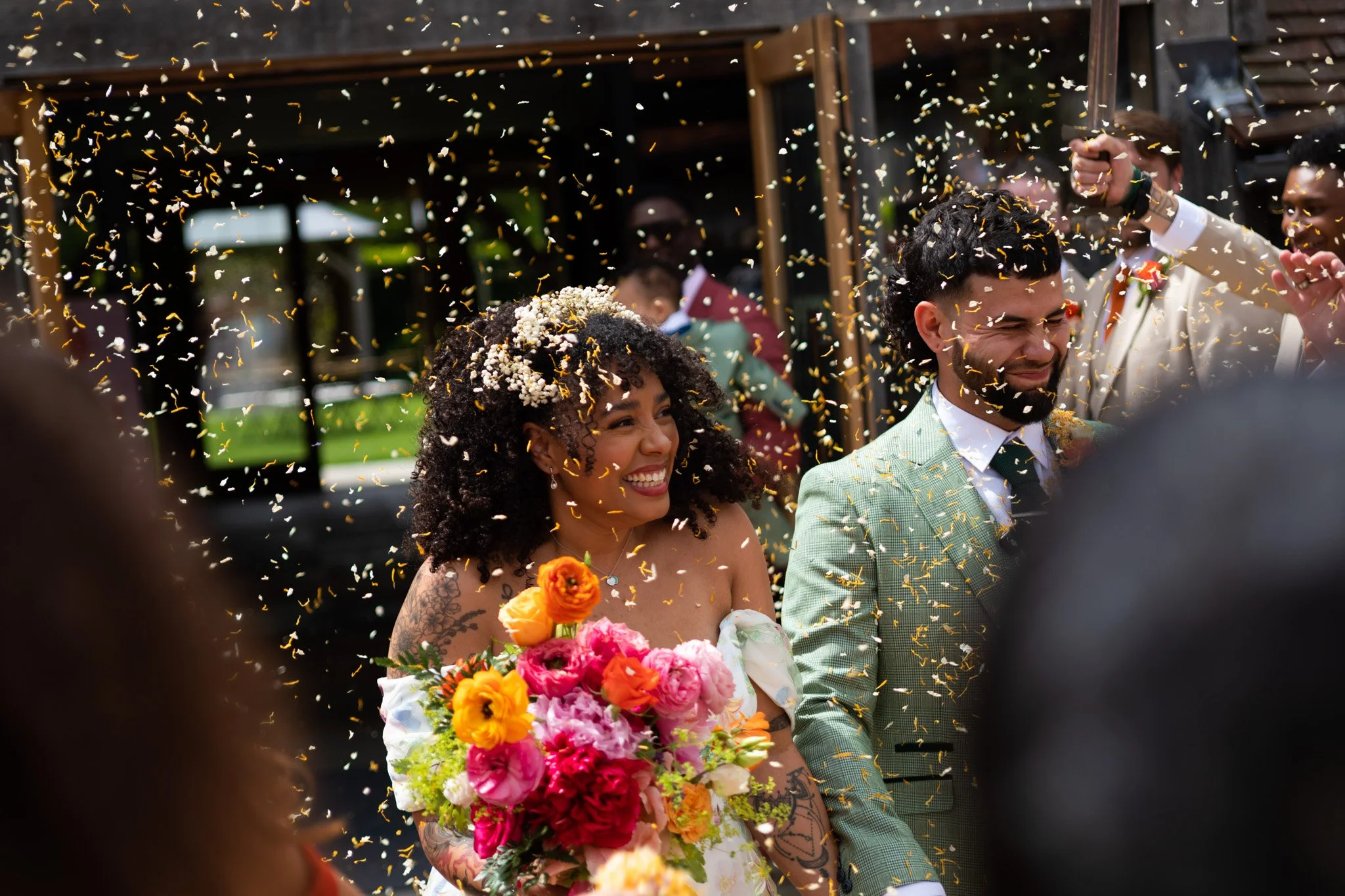 A smiling woman with curly hair holding a large bouquet of colorful flowers and wearing a white dress, and a man in a green plaid suit celebrating under falling confetti at a wedding.