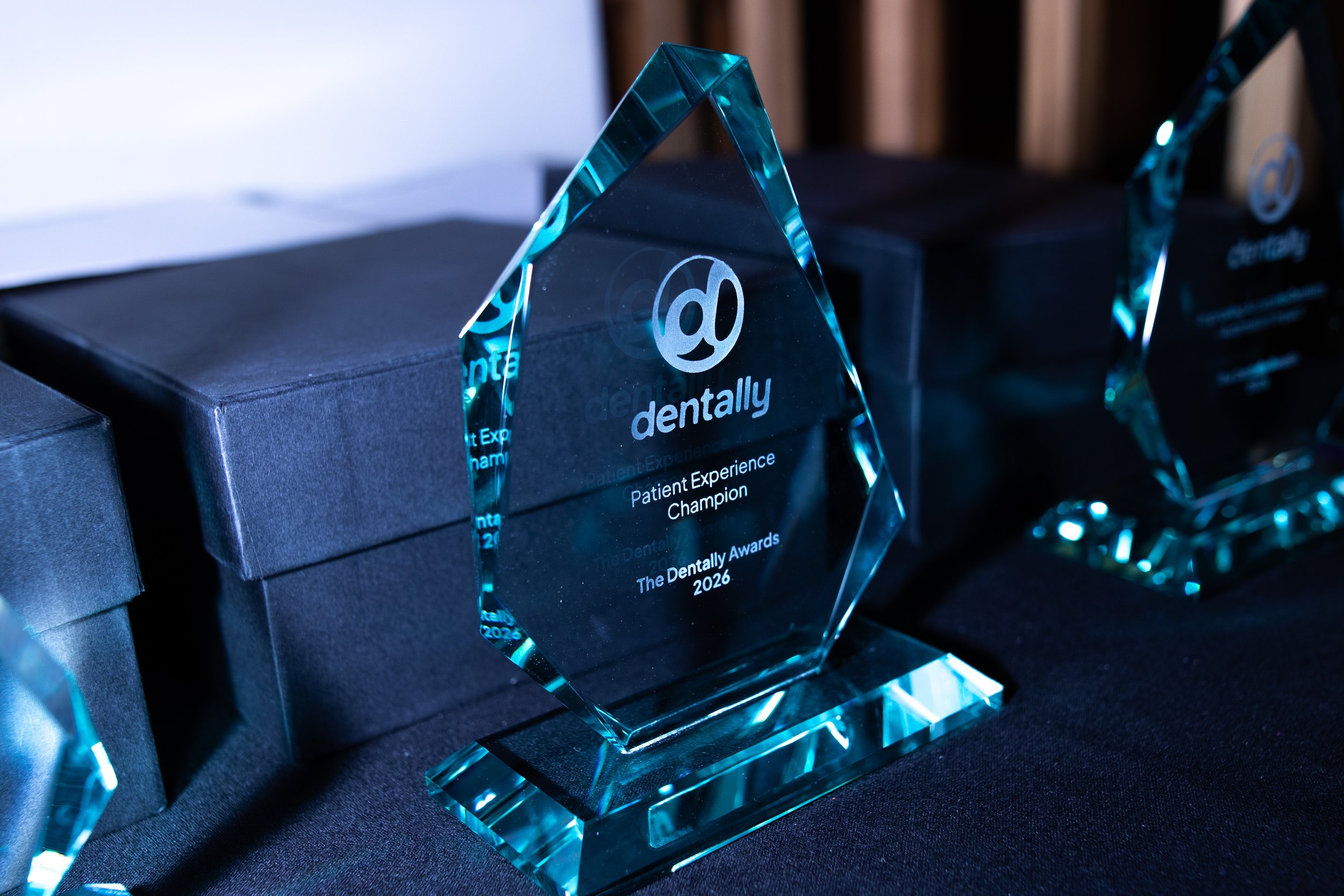 A glass award shaped like a pyramid with the Dentally logo and text engraved on it, displayed on a black surface with other awards in the background.