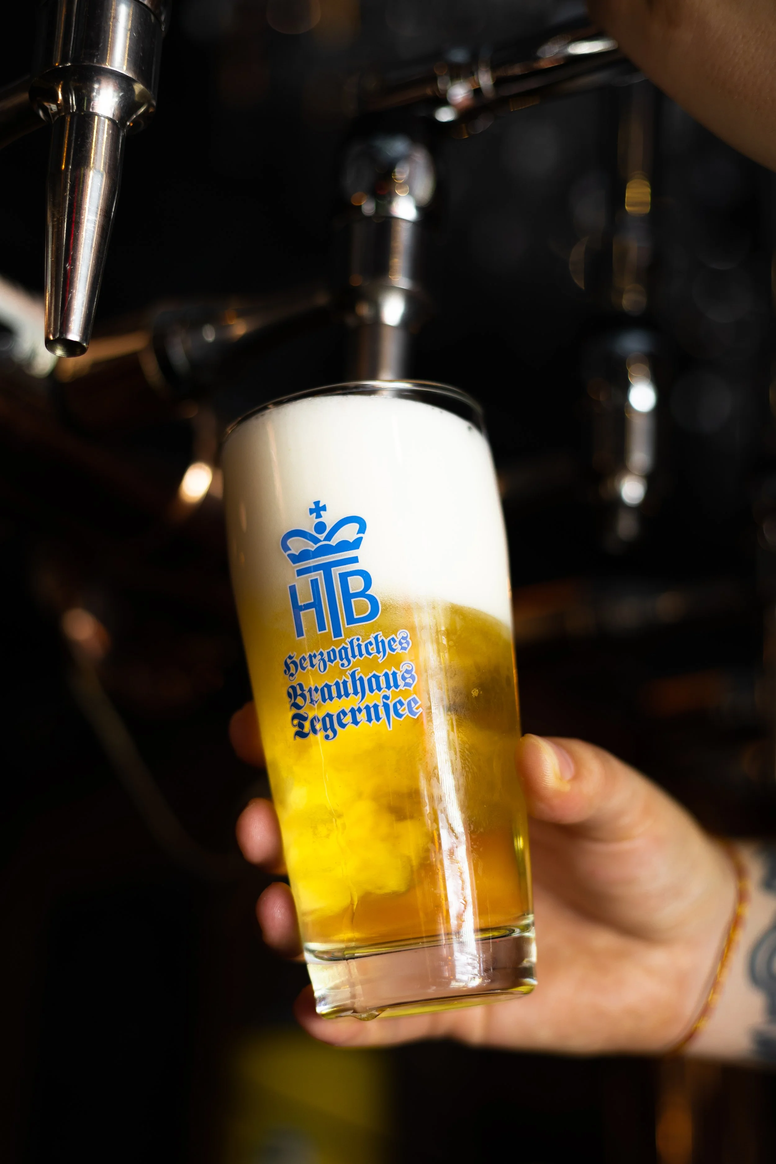 A hand holding a tall glass of beer under a beer tap. The beer glass has a logo with a crown and the words "Herzogliches Brauhaus Tegernsee" on it.