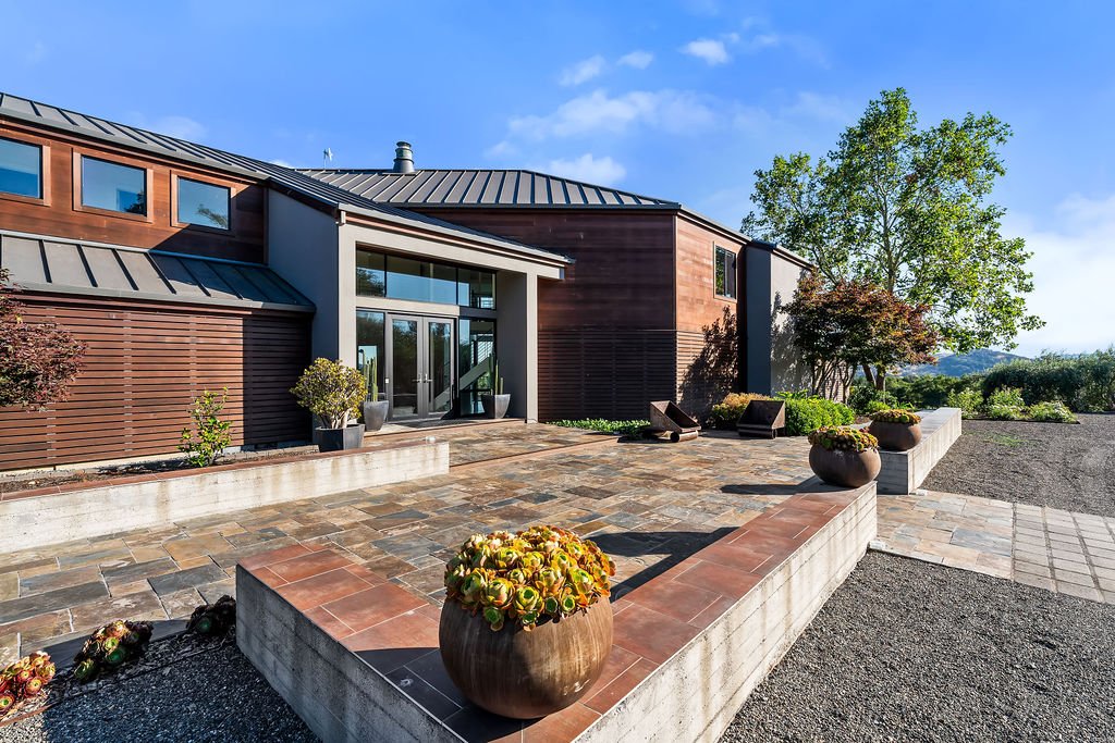 Modern house with a stone patio, potted plants, and outdoor seating, surrounded by trees and greenery under a blue sky.
