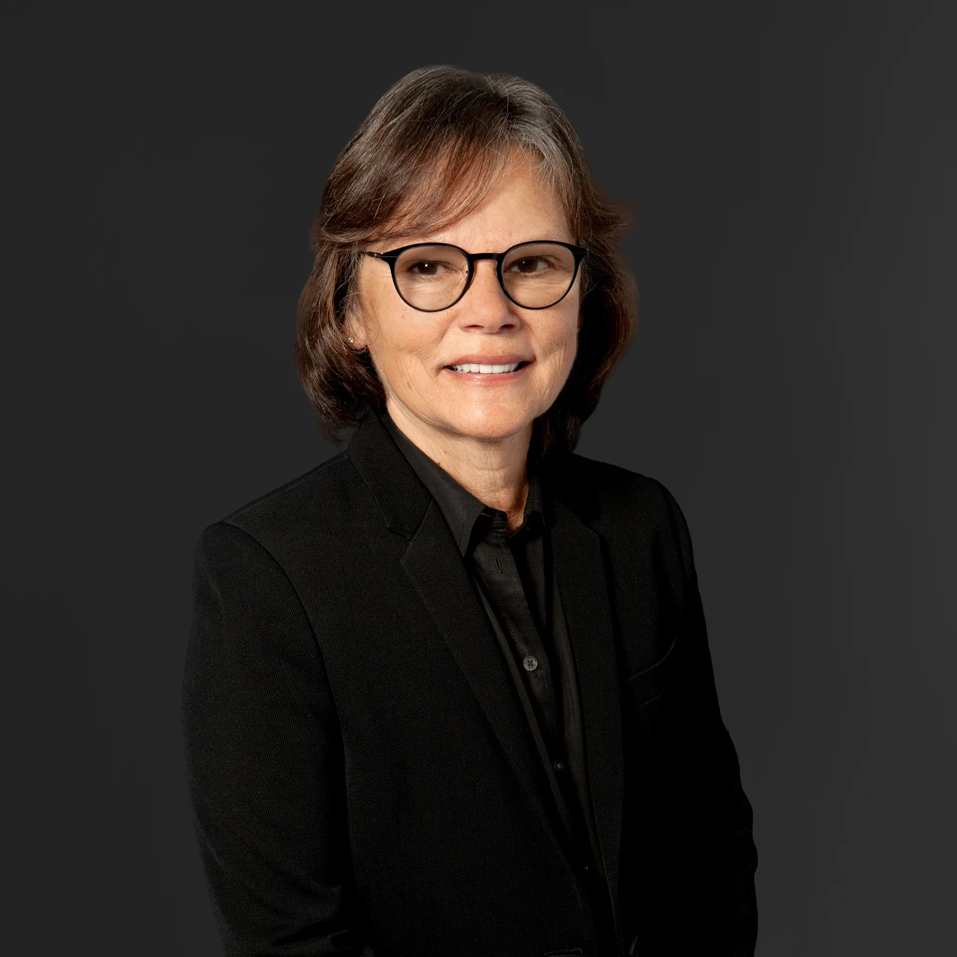A portrait of a middle-aged woman with short brown hair, wearing glasses, and dressed in a black suit against a dark background.