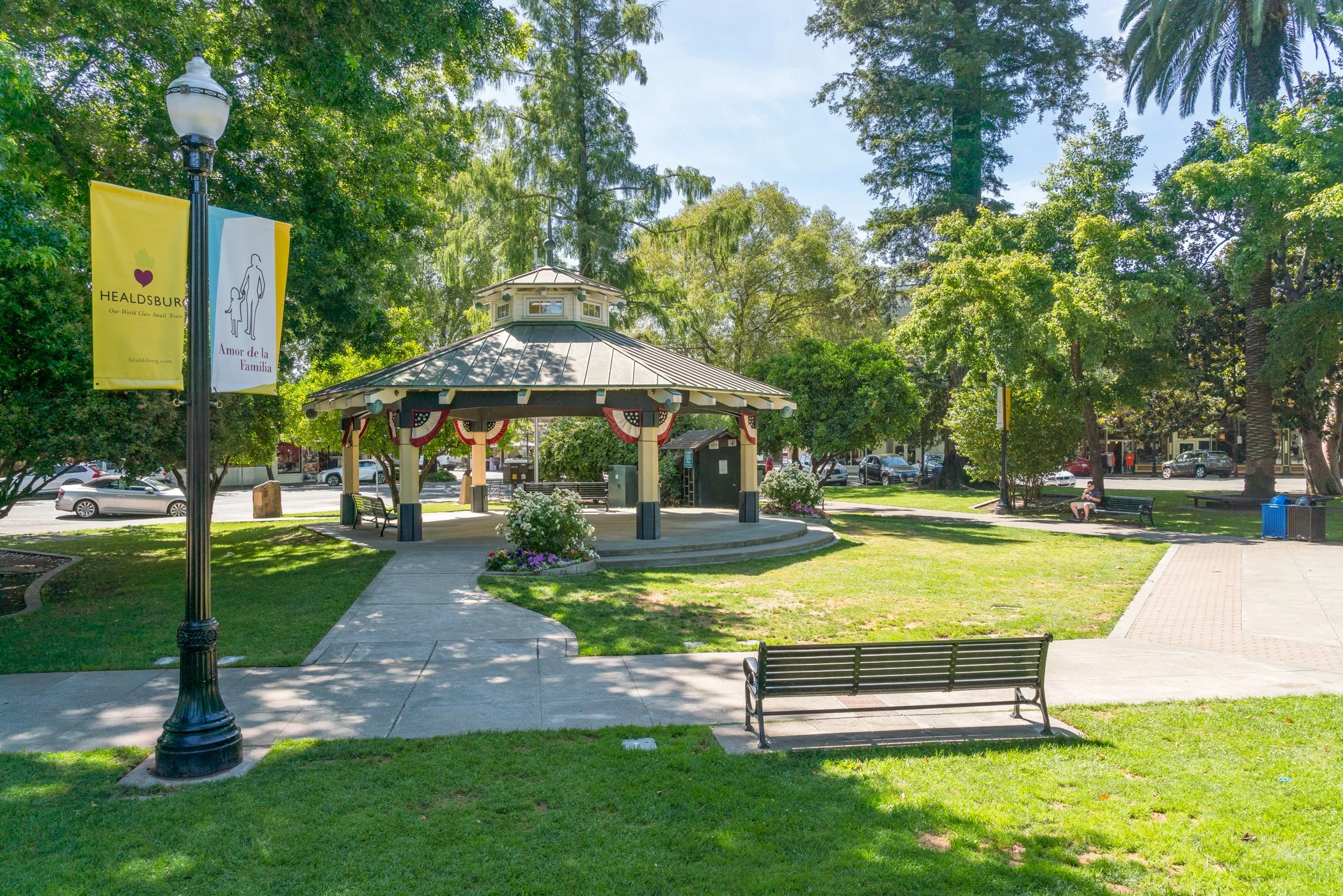A small park with a gazebo decorated with red, white, and blue bunting, surrounded by green trees and grass, with benches, lampposts, and parked cars visible in the background.