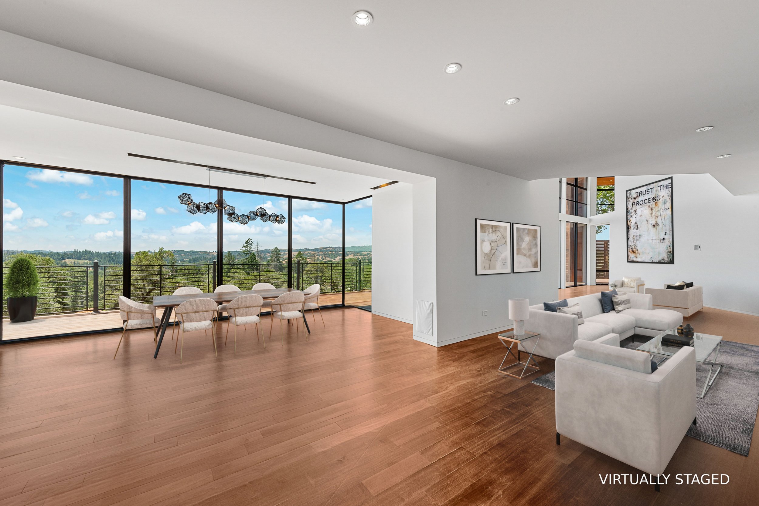 Living room and dining area with large windows showing an outdoor balcony and scenic view, white walls, wooden flooring, modern furniture, and decorative art.