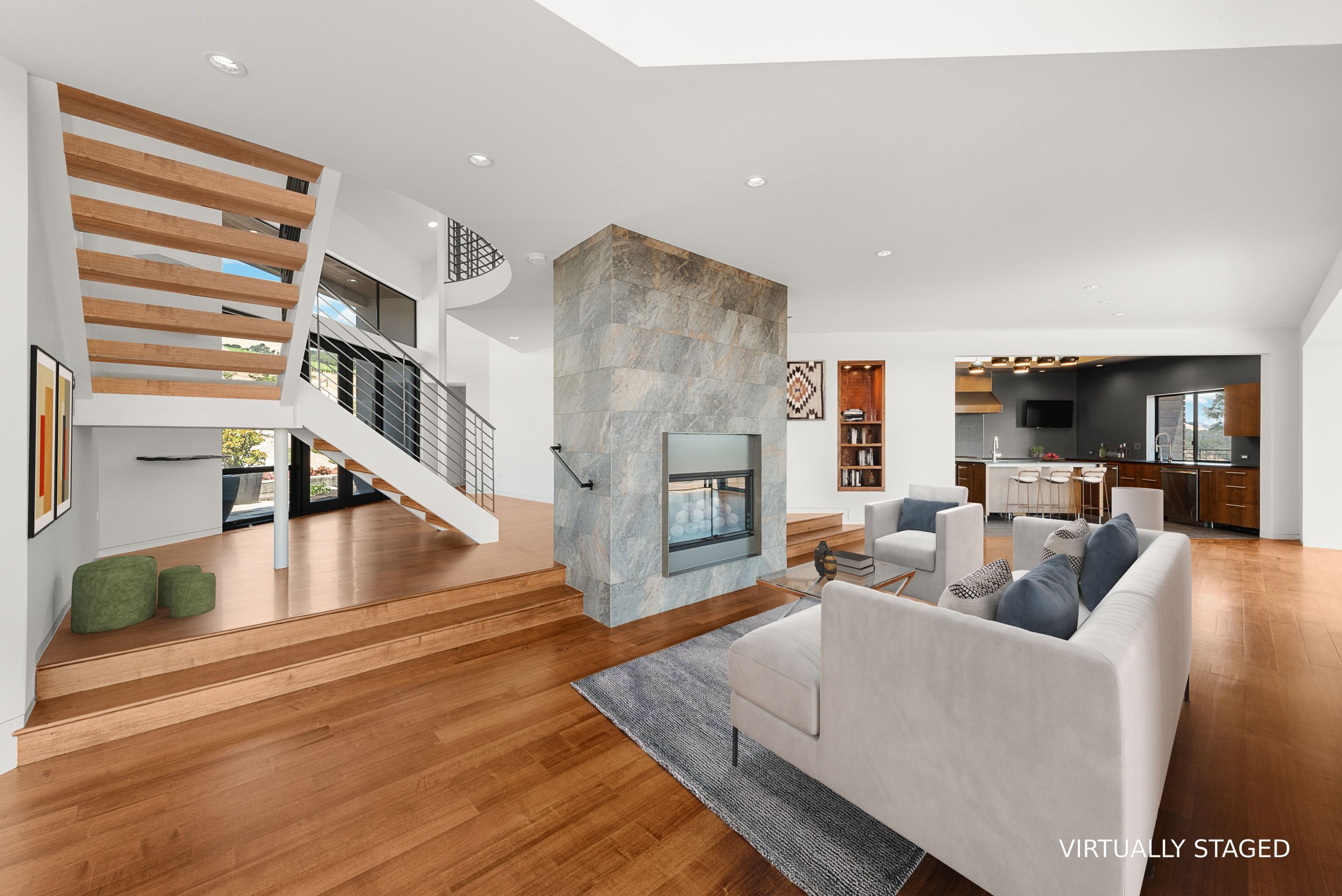 Interior of a modern, open-concept living space with a staircase, stone fireplace, and kitchen area with bar stools. The space has wood floors, white walls, and large windows.