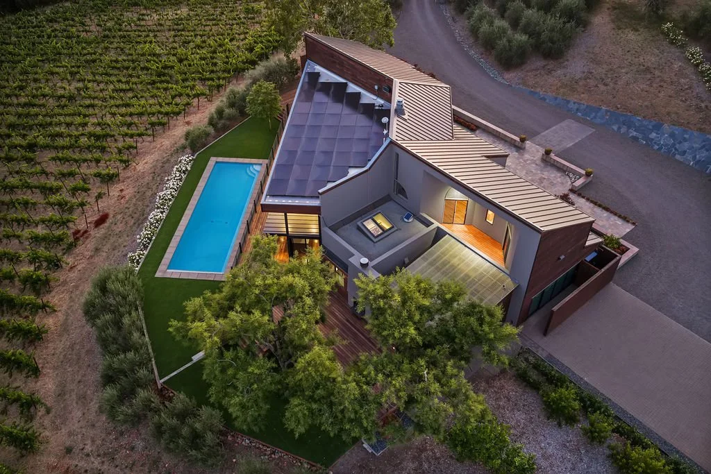 Aerial view of a modern house with a backyard pool, surrounded by trees and situated next to vineyard rows, with a paved driveway, on a partly cloudy day.
