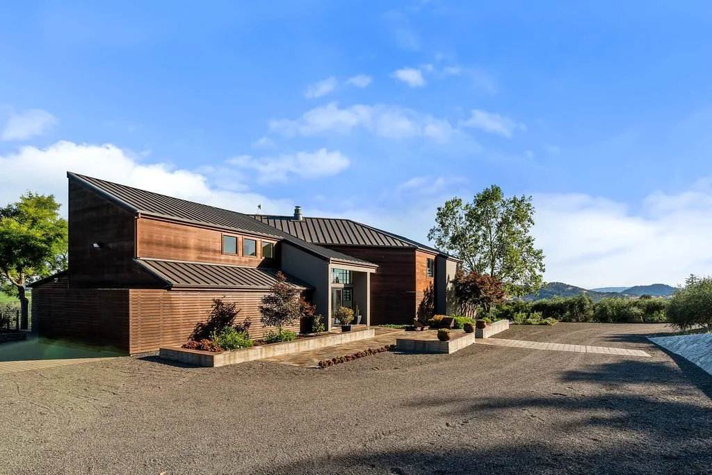 Modern house with wood siding and metal roof, surrounded by landscaped greenery and open outdoor space, under a blue sky with clouds.