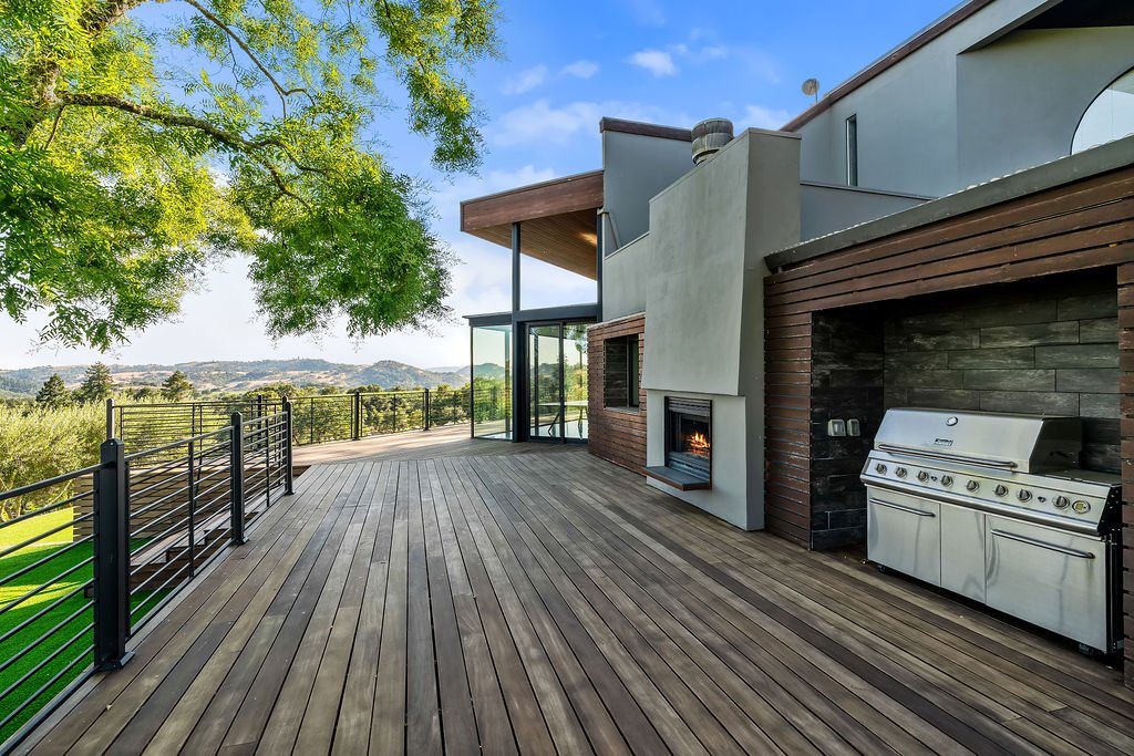 Modern house outdoor deck with grilling area, fireplace, glass doors, and scenic view of distant hills and greenery.