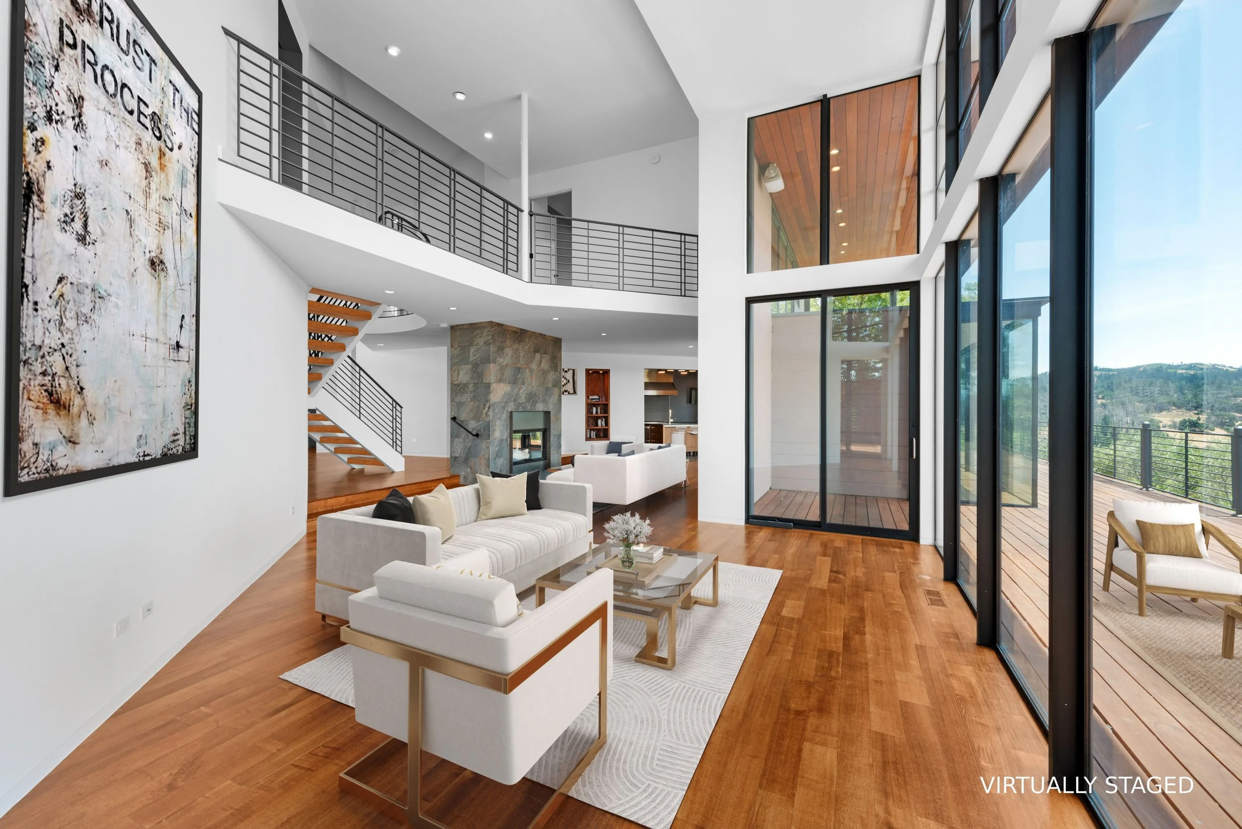 Modern living room with white sofas, wooden floors, large glass windows, and a balcony view of lush green hills.