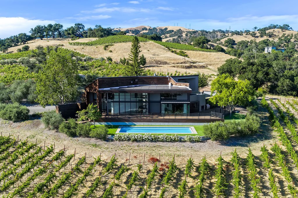 A modern house with large glass windows is surrounded by vineyards and trees in a hilly landscape, with a swimming pool in the backyard.