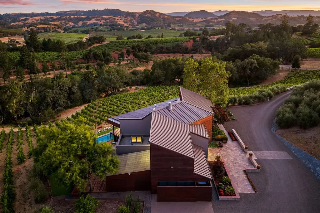 A modern house with metal roofing and a backyard swimming pool, situated in a rural landscape with vineyards and rolling hills at sunset.