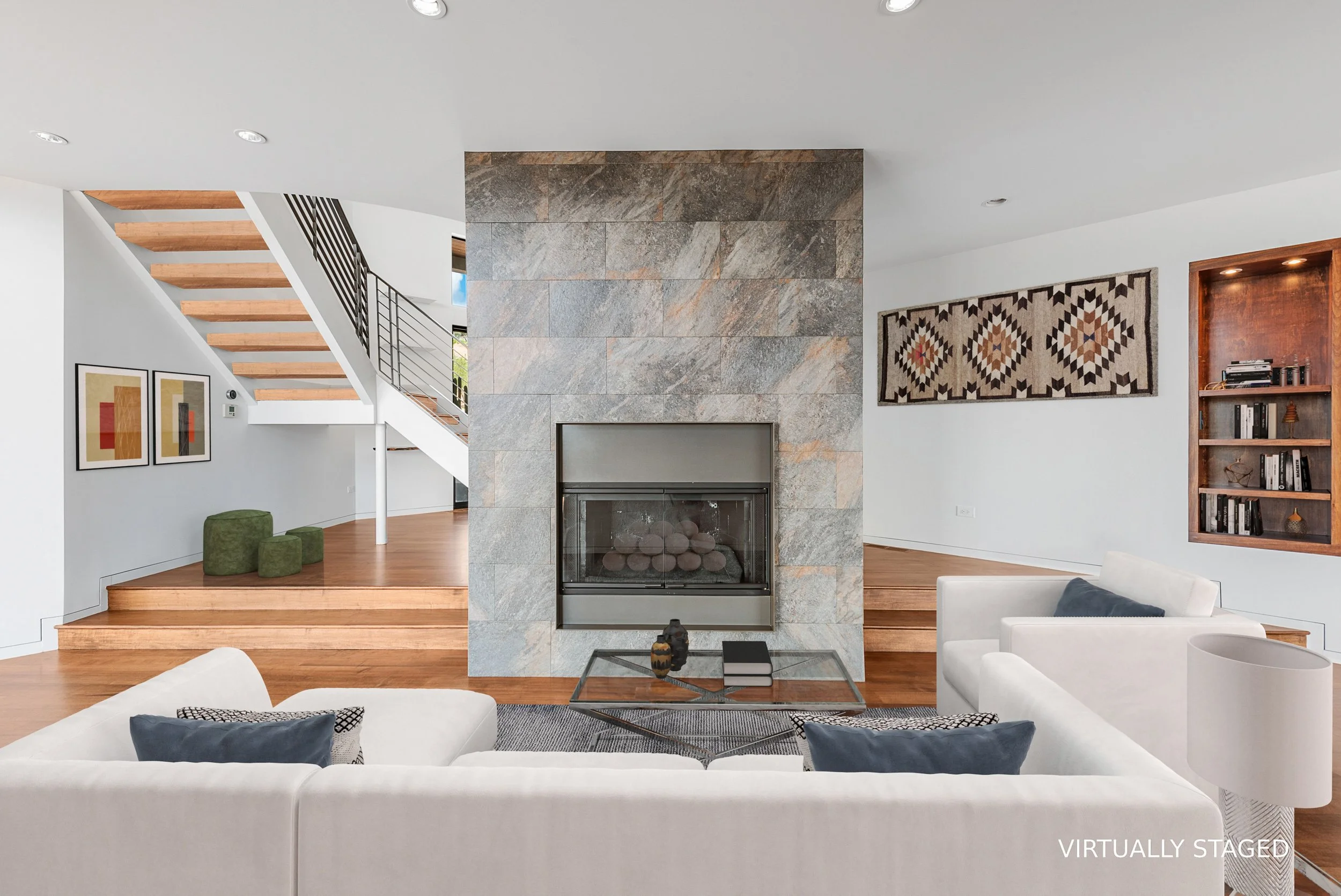 Modern living room with stone fireplace, white couches with blue and black pillows, wooden floors, and artwork on the walls.