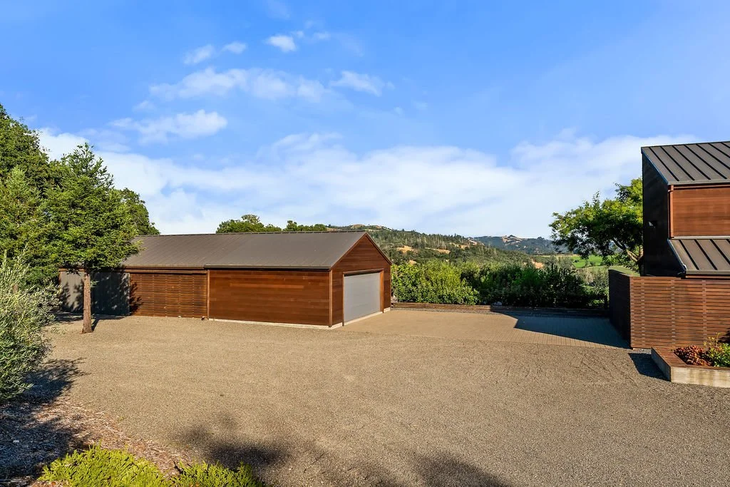 A modern house with wooden siding and a metal roof, set in a rural landscape with gravel driveway and greenery, under a partly cloudy sky.