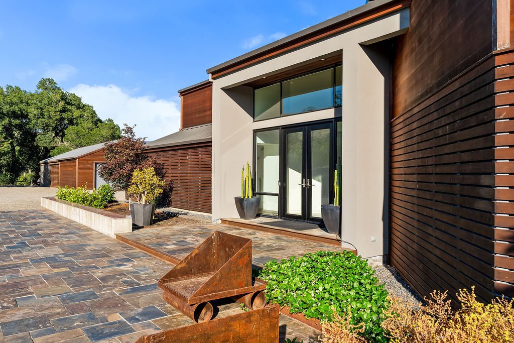 Modern building with large glass entry doors, wooden siding, plants in pots, and a paved outdoor area.