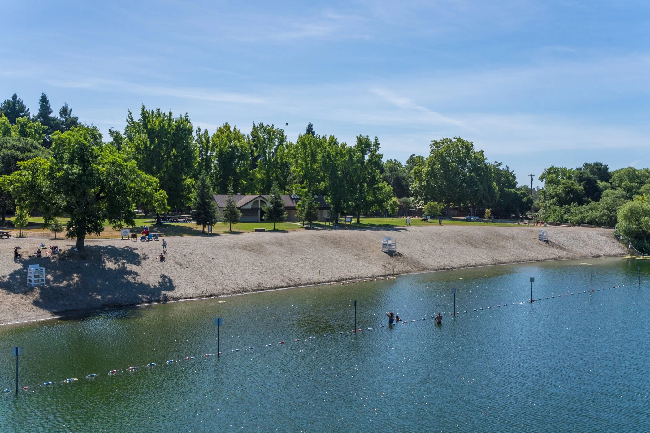 A park area with a sandy shore along a river or lake, with people swimming and relaxing near the water, surrounded by green trees and a clear blue sky.