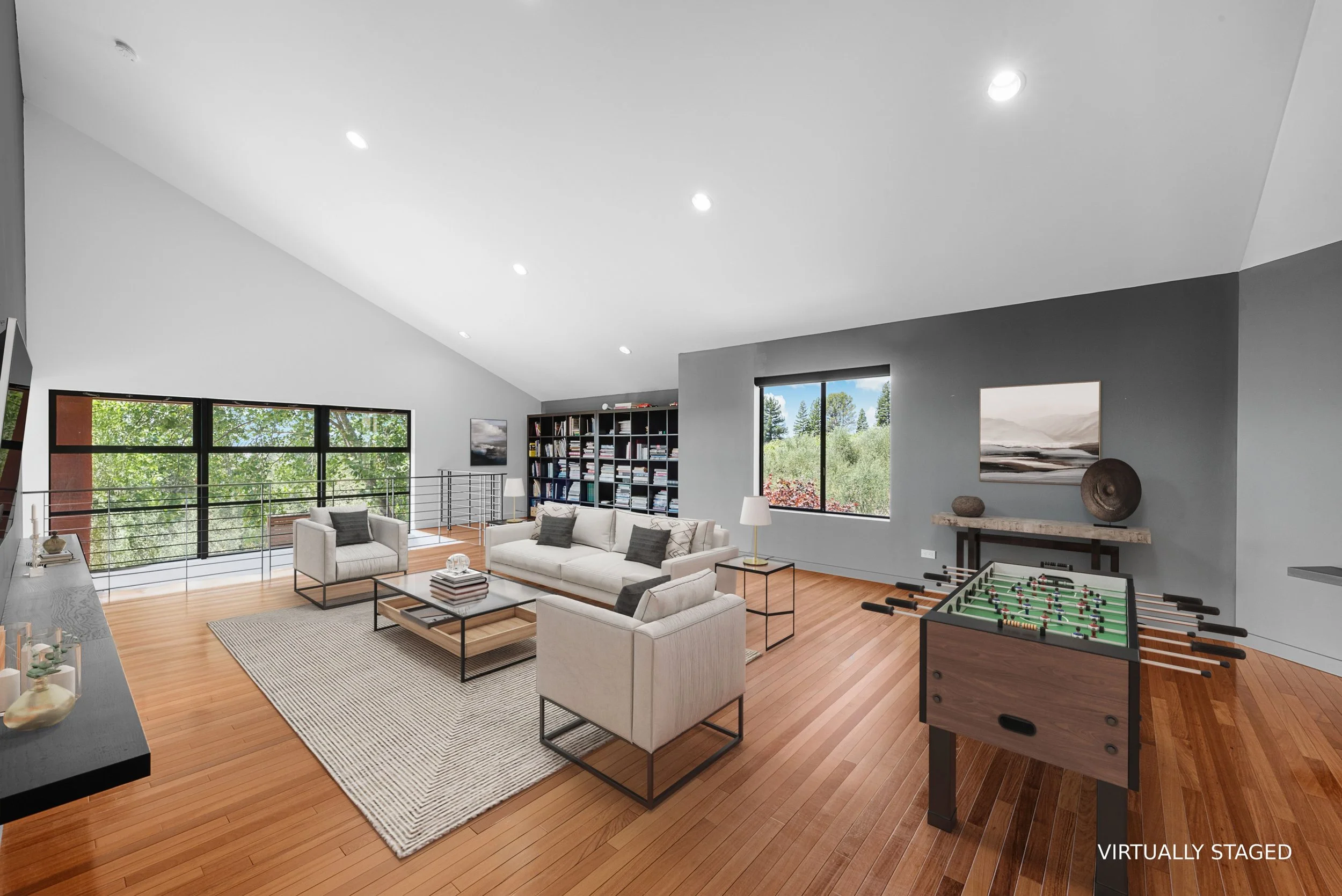 Living room with white furniture, wooden floor, large windows showing trees outside, bookshelves, wall art, foosball table, and modern decor.