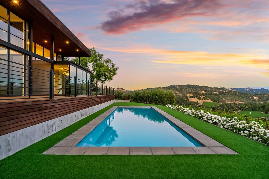 Modern house with large glass windows and a wooden balcony, overlooking a backyard with a rectangular swimming pool, green artificial grass, white flowers, lush hills, and a colorful sunset sky.
