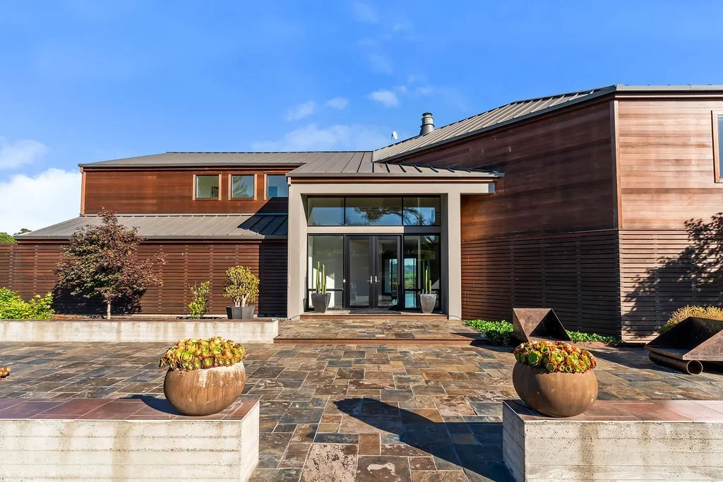 Modern house with wooden exterior, glass entrance, and stone patio under a blue sky.