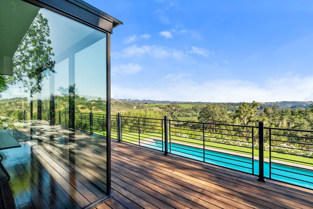 A modern outdoor deck with wooden flooring and a glass wall overlooking a green landscape and blue sky, with a swimming pool visible below.