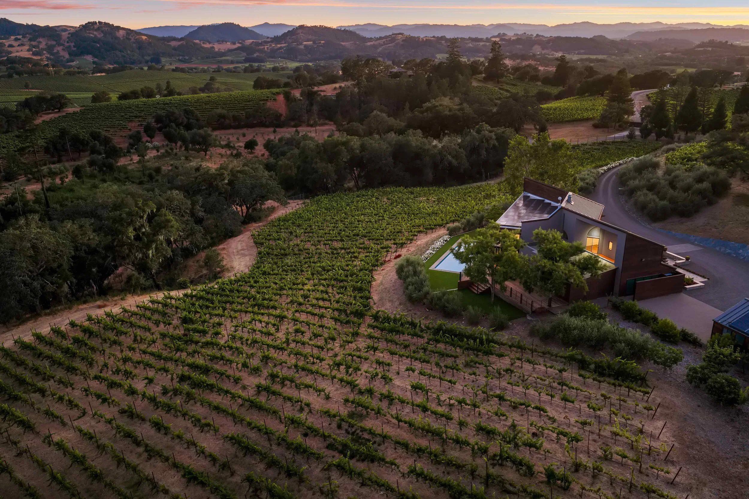 A scenic view of rolling hills with lush vineyards, trees, and a modern house with a swimming pool at sunset.