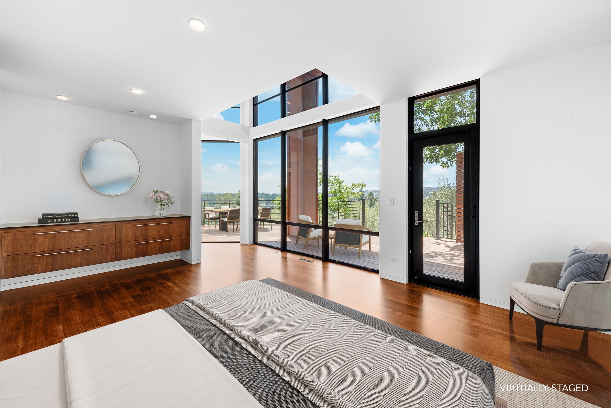 Interior of a modern apartment living room with white walls, hardwood floors, large glass sliding doors leading to a balcony with outdoor furniture, and a round mirror on the wall. Bright natural light and a view of green trees and blue sky outside.