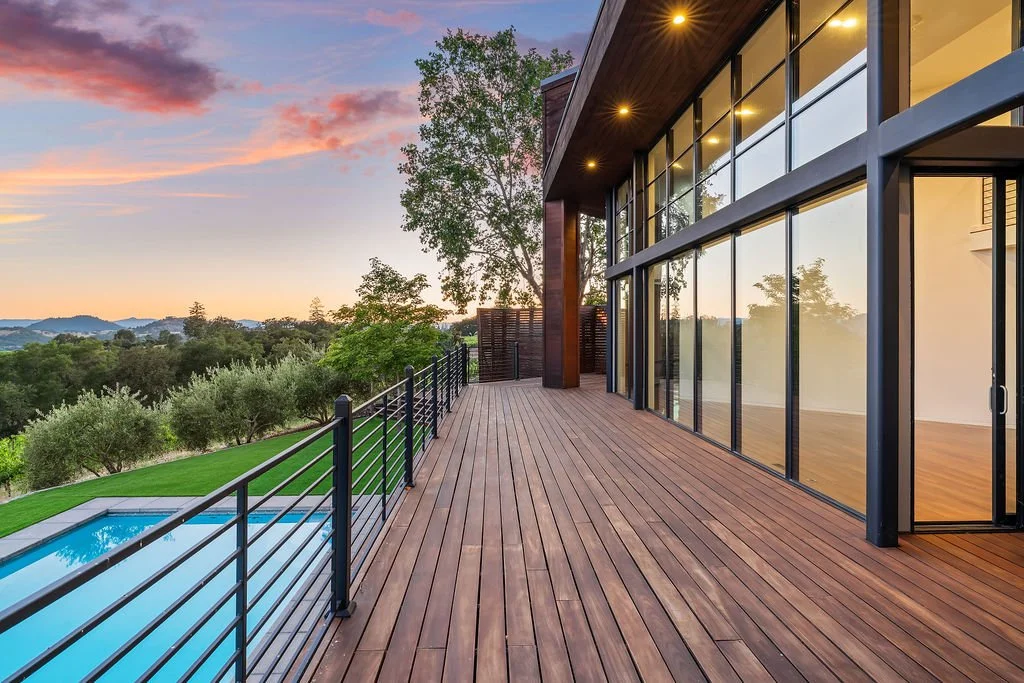 Modern house with large glass windows and wooden deck overlooking a landscape with trees, a pool, and a sunset sky.