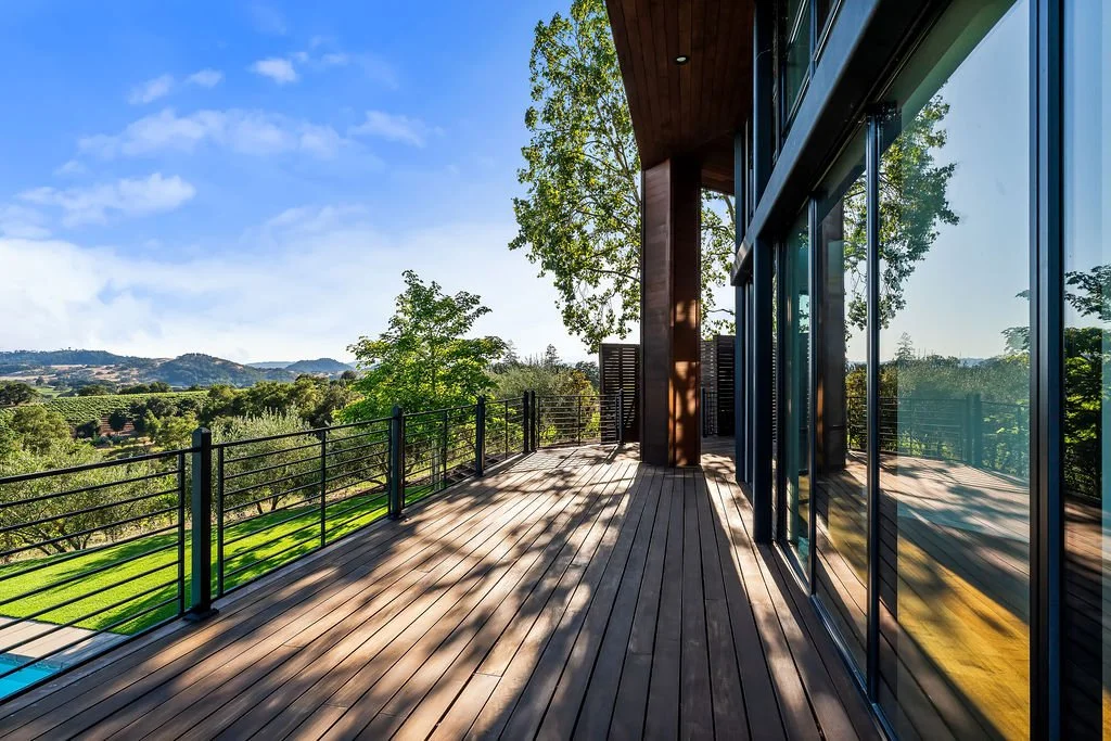 Modern house balcony with wooden floor, glass sliding doors, and a view of trees and hills under a blue sky.