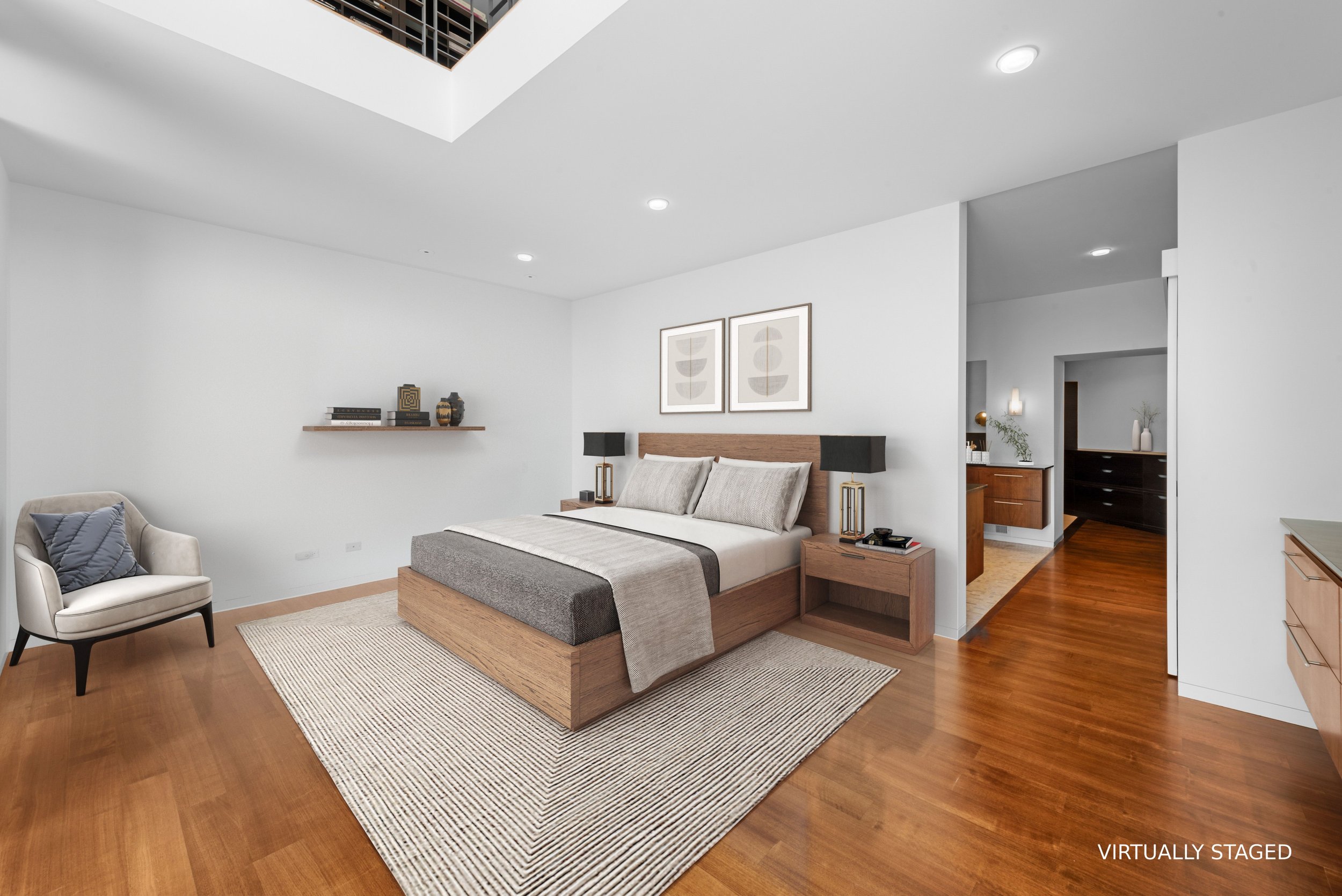 A bedroom with a wooden bed, two black lamps, framed art above the bed, a side table, a beige armchair with a blue pillow, and a wooden shelf with decorative objects, against white walls and wooden flooring.