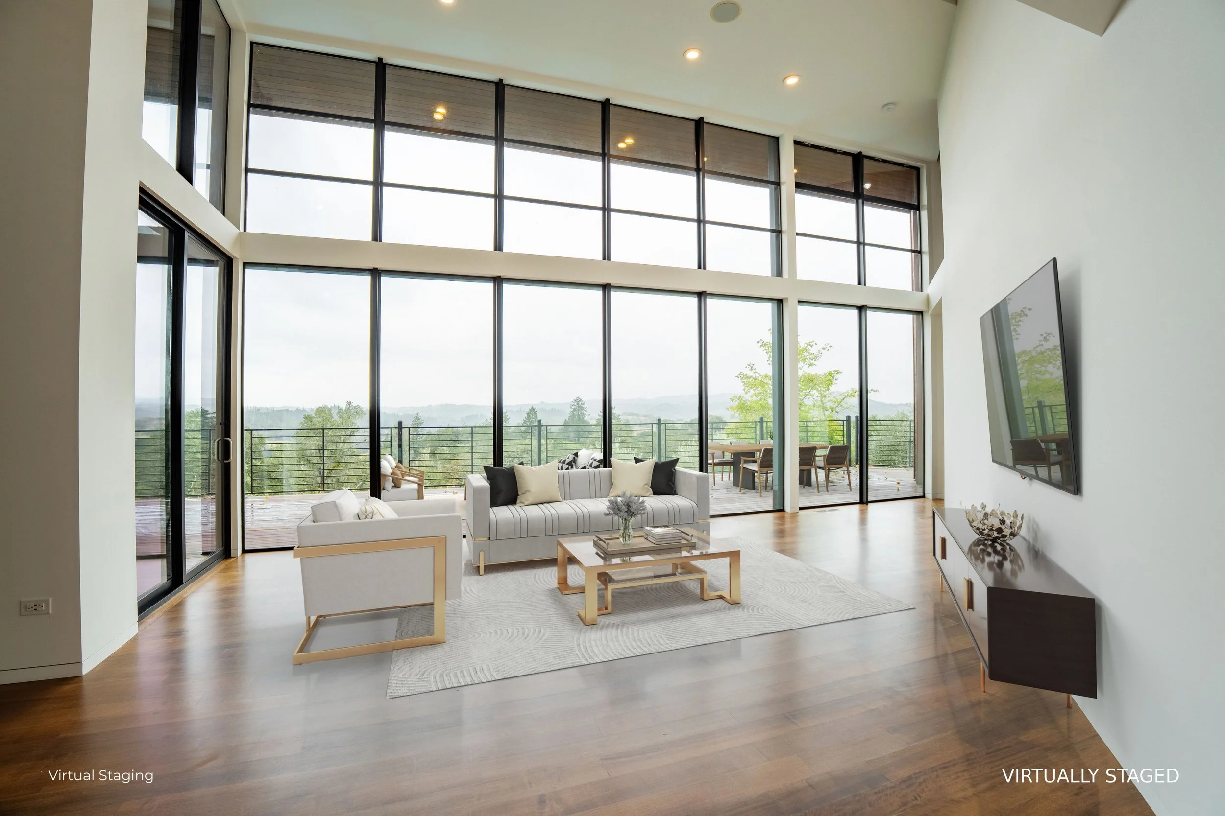 A spacious modern living room with large floor-to-ceiling windows facing a balcony with a view of trees and hills in the distance. The room features a white sofa with black and white pillows, a wooden coffee table with books and a small decorative it