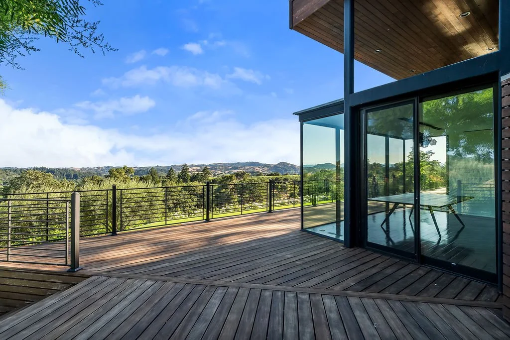 Modern house with a large wooden deck and glass walls, overlooking a green landscape with trees and hills under a blue sky.