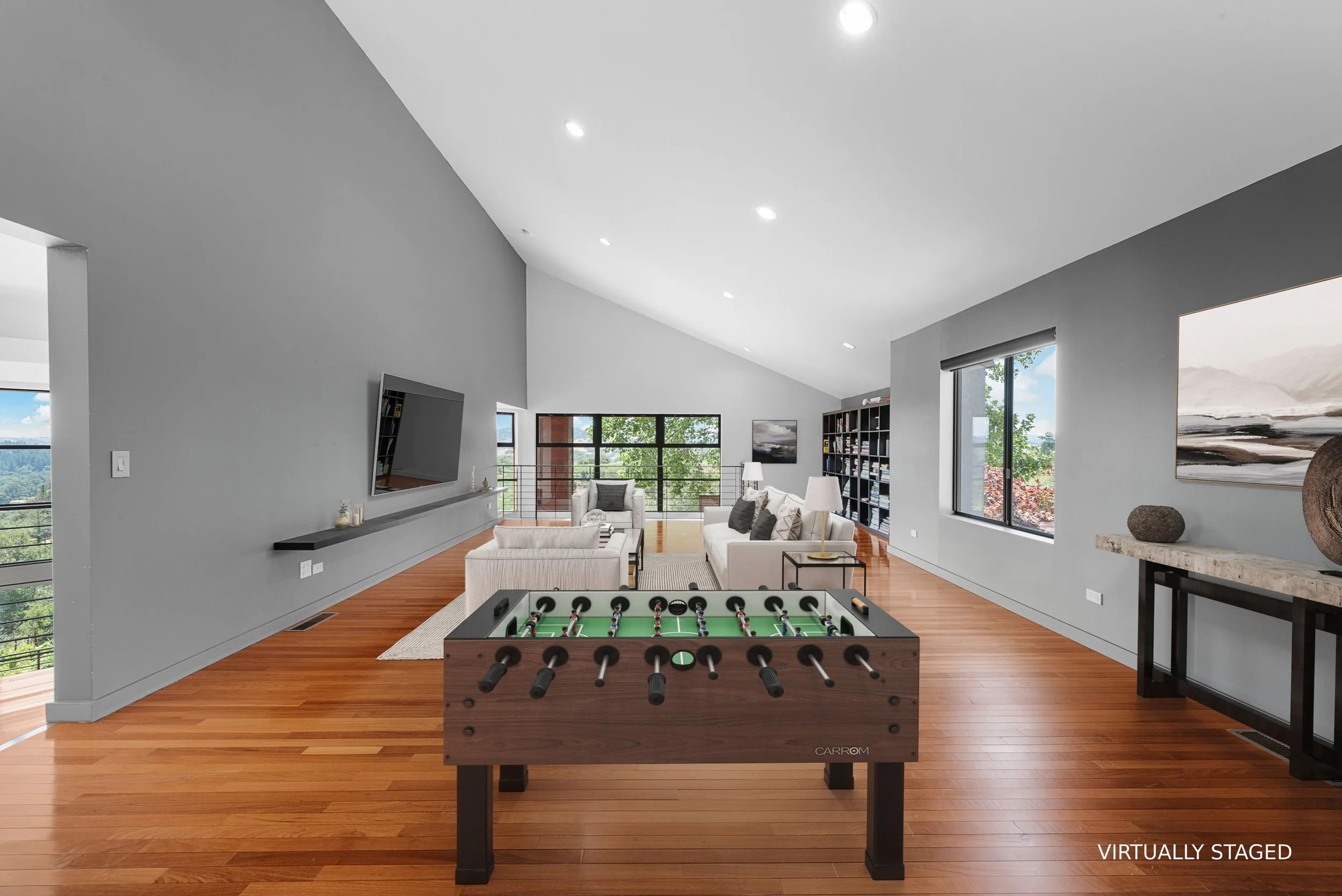 A modern living room with hardwood floors, gray walls, and large windows showing trees outside. The room has a white sofa, an armchair, a TV mounted on the wall, and a bookshelf. A foosball table is in the foreground, with the words 'VIRTUALLY STAGED