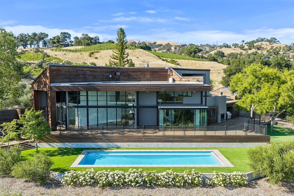 Modern house with large glass windows, a wooden deck, a swimming pool, and scenic hillside landscape in the background.