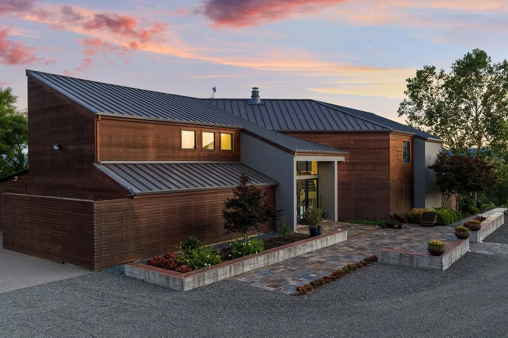 Modern house with metal roof and wooden siding, surrounded by landscaped garden and trees at sunset.