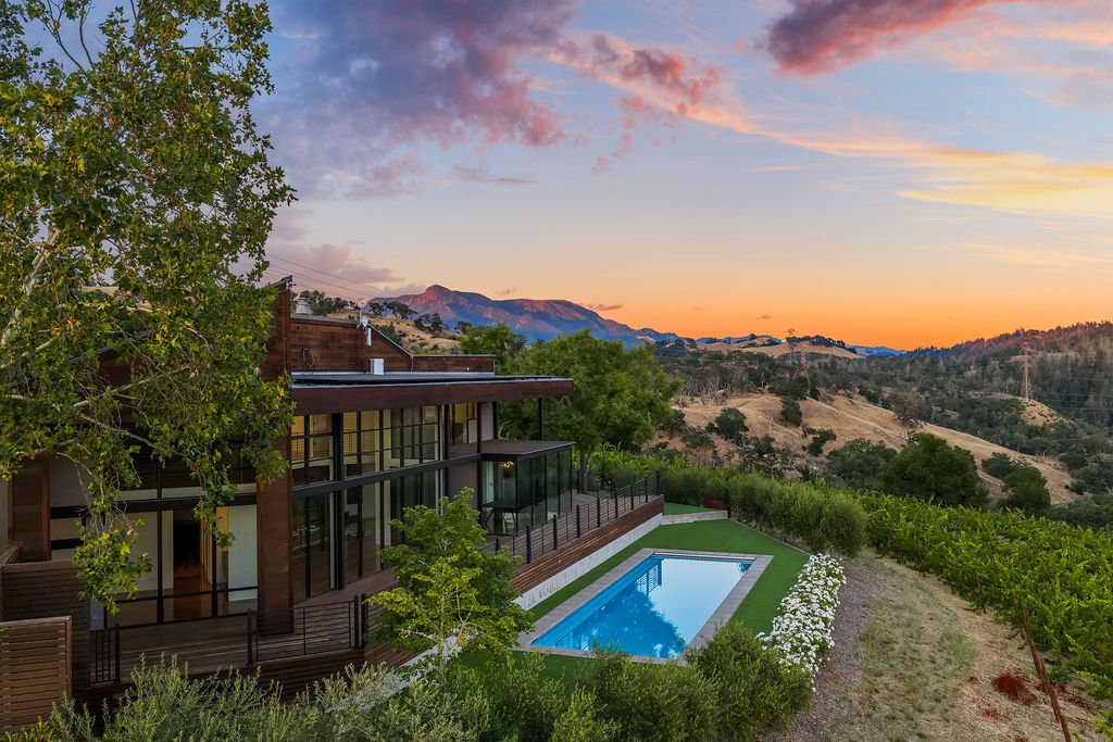 Modern house with large windows and a backyard with a swimming pool, hillside landscape, and mountains at sunset.